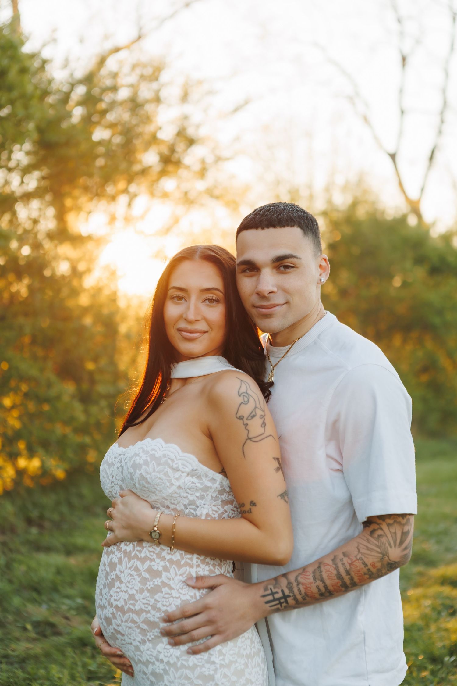 Pregnant couple embracing at sunset outdoors, woman in white lace dress, man with tattoos hugging her baby bump.