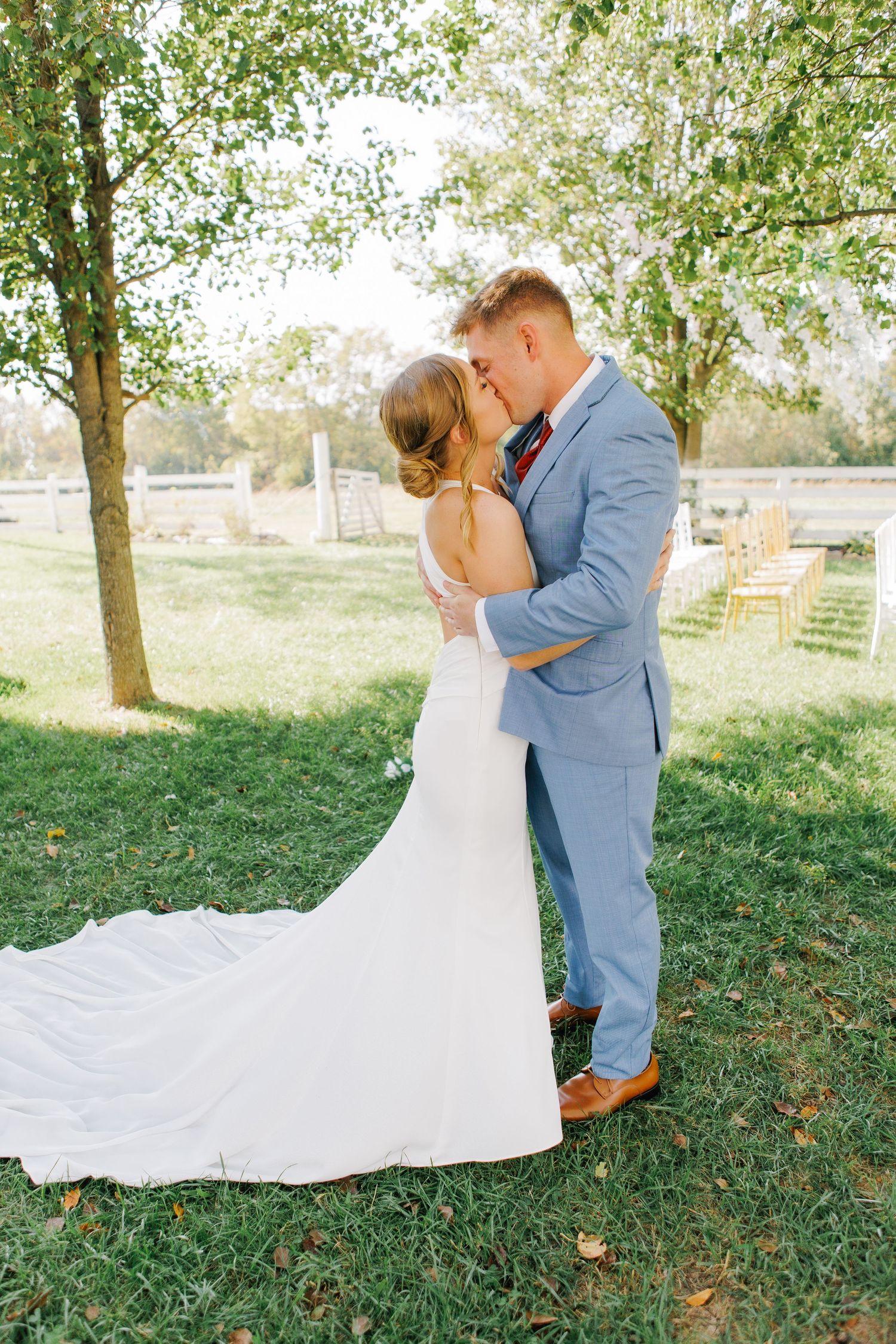 Bride and groom share a first kiss on green grass outdoors, surrounded by trees at their elegant wedding ceremony.