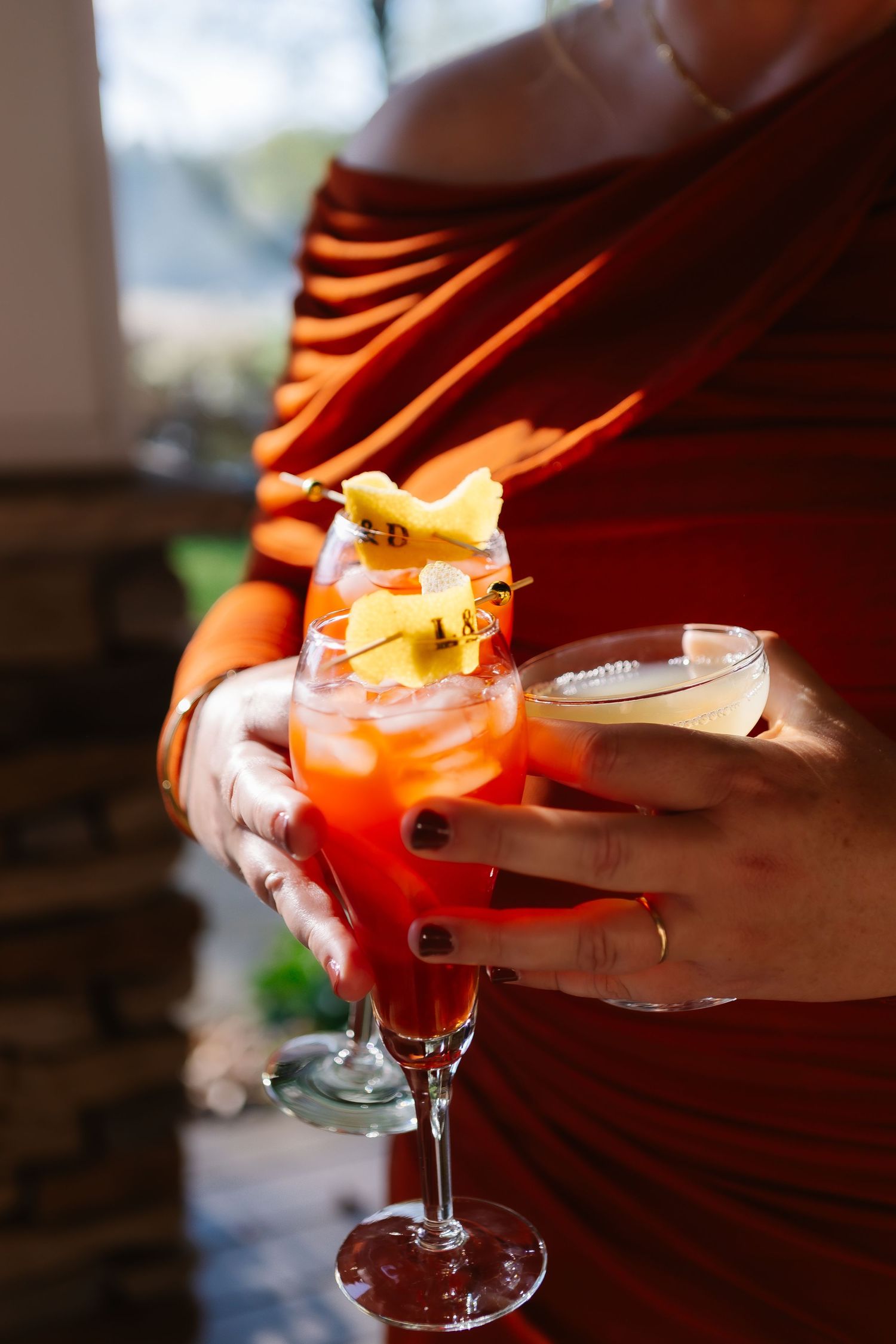 A hand holds a vibrant red cocktail garnished with citrus in a coupe glass against a warm-lit background.