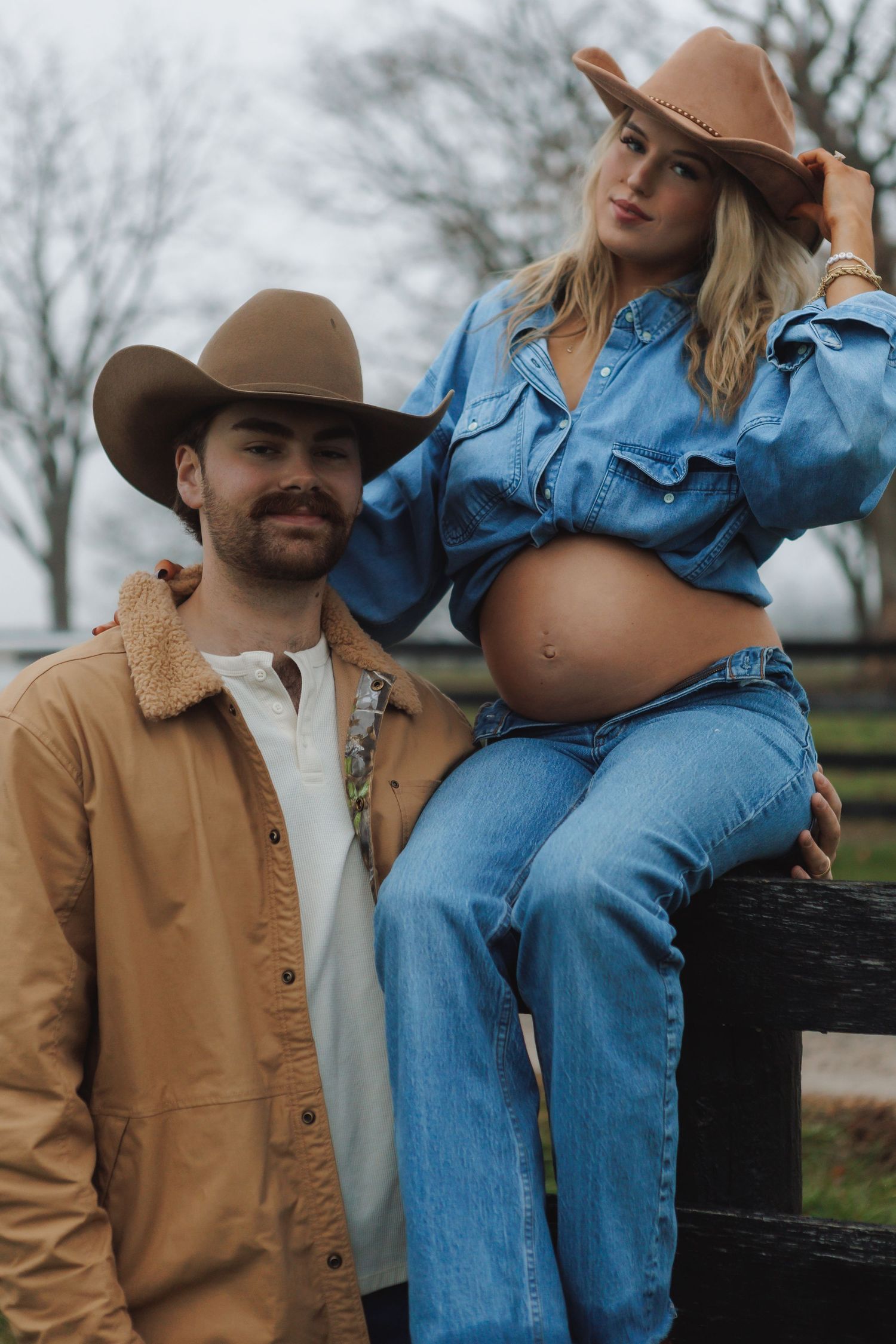Couple in western wear sits on black wooden fence during overcast day.