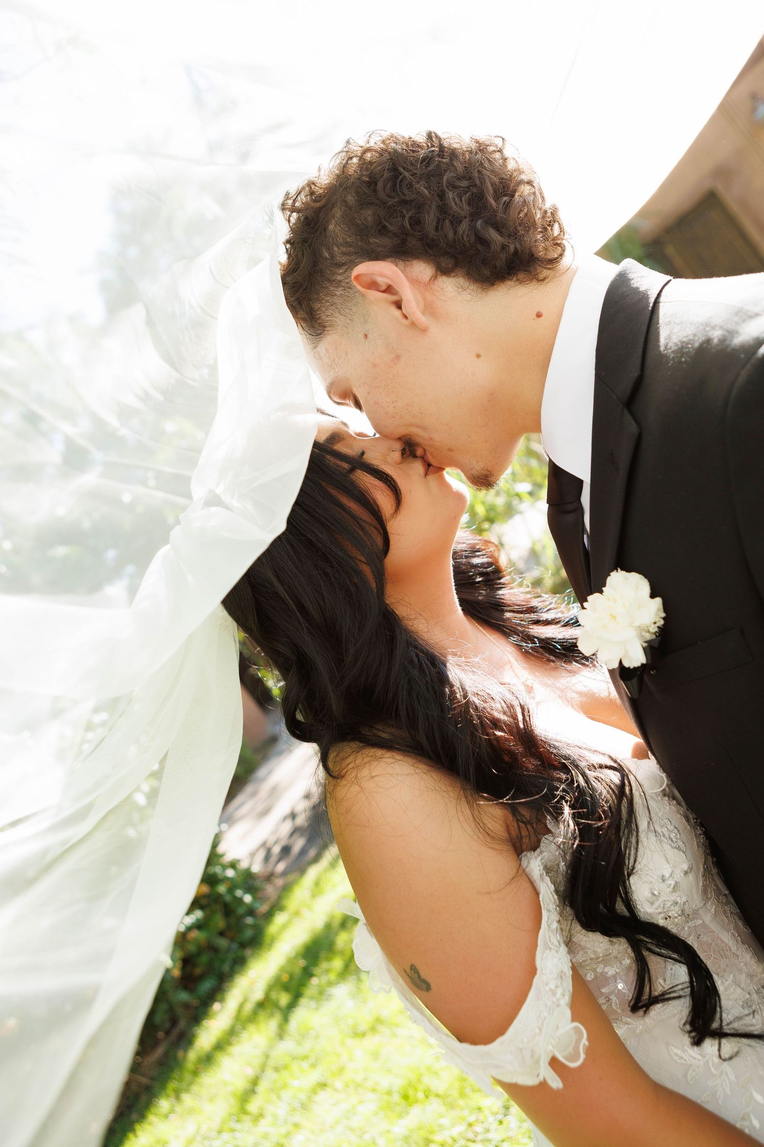 A bride and groom share a romantic kiss outdoors on their wedding day, with the bridal veil flowing in the breeze.