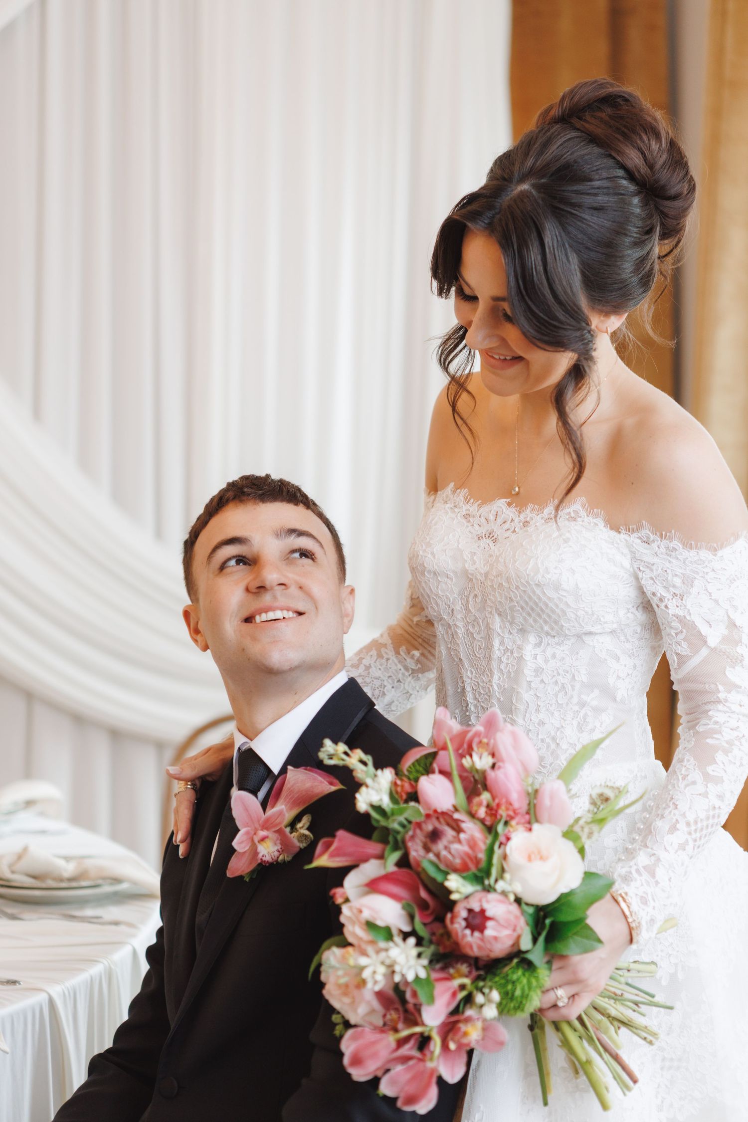 Smiling groom in black suit looks up at bride in lace off-shoulder wedding dress holding pink floral bouquet.