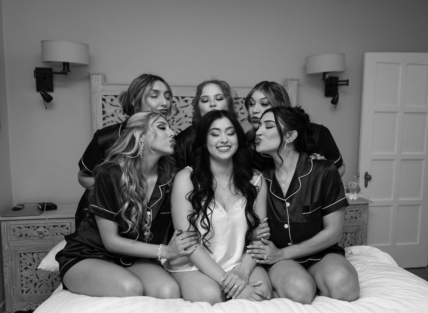 Black and white photo of a bride surrounded by bridesmaids in satin pajamas sharing kisses on a bed.