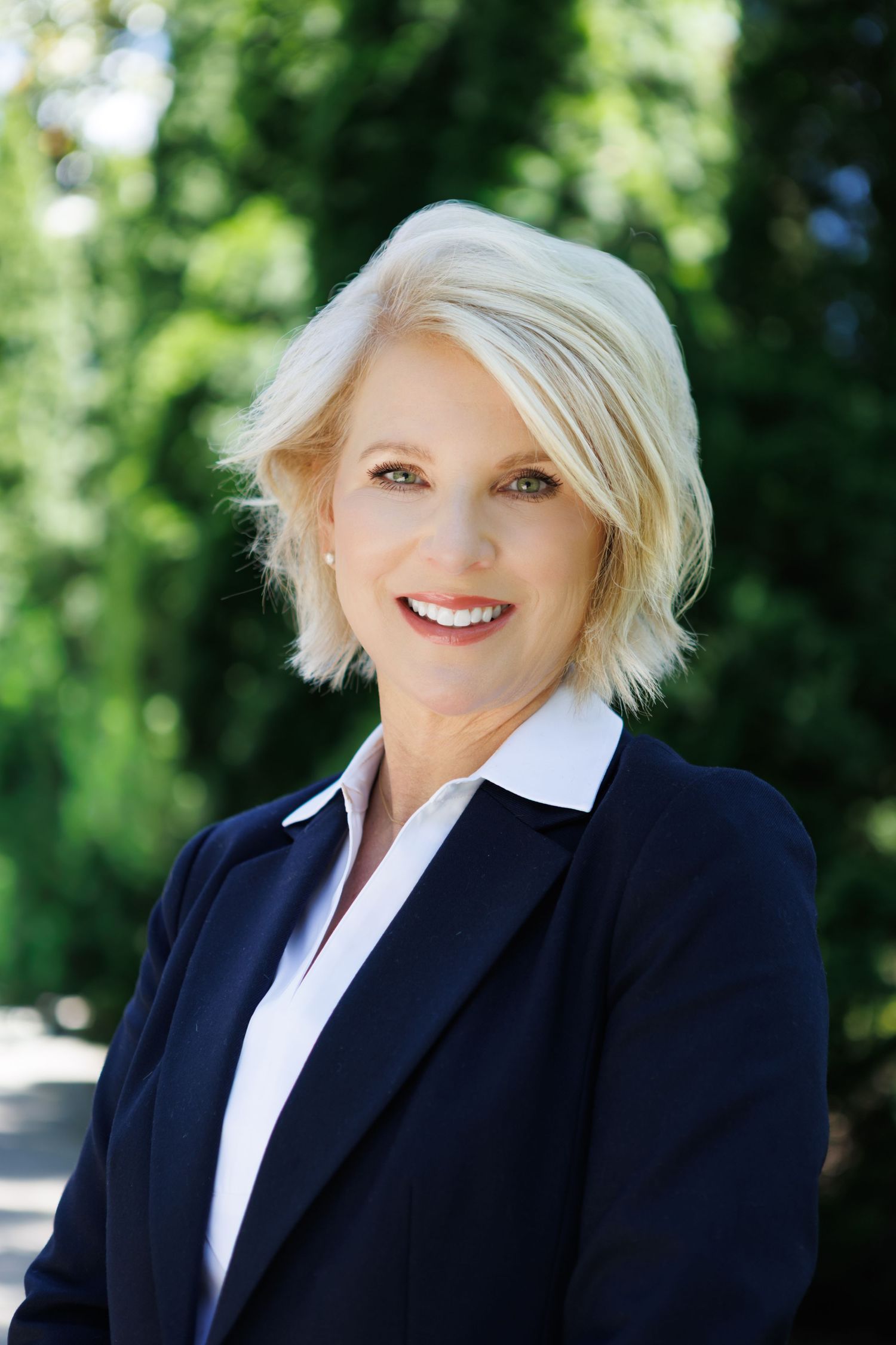 Professional headshot of a smiling blonde woman in a navy blazer outdoors with green trees in the background.