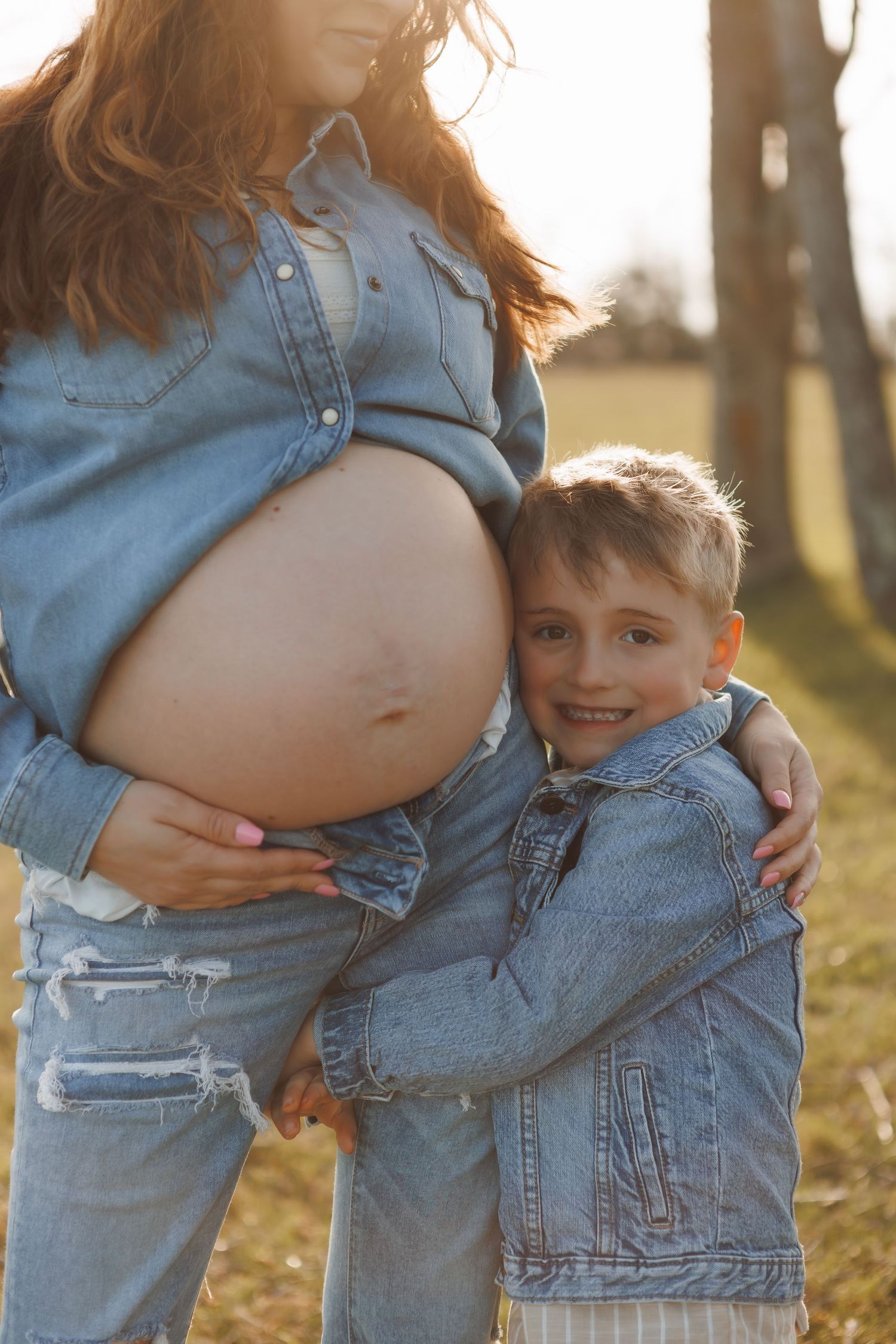 A young boy in a denim jacket hugs his pregnant mother outdoors, smiling warmly in golden sunlight.