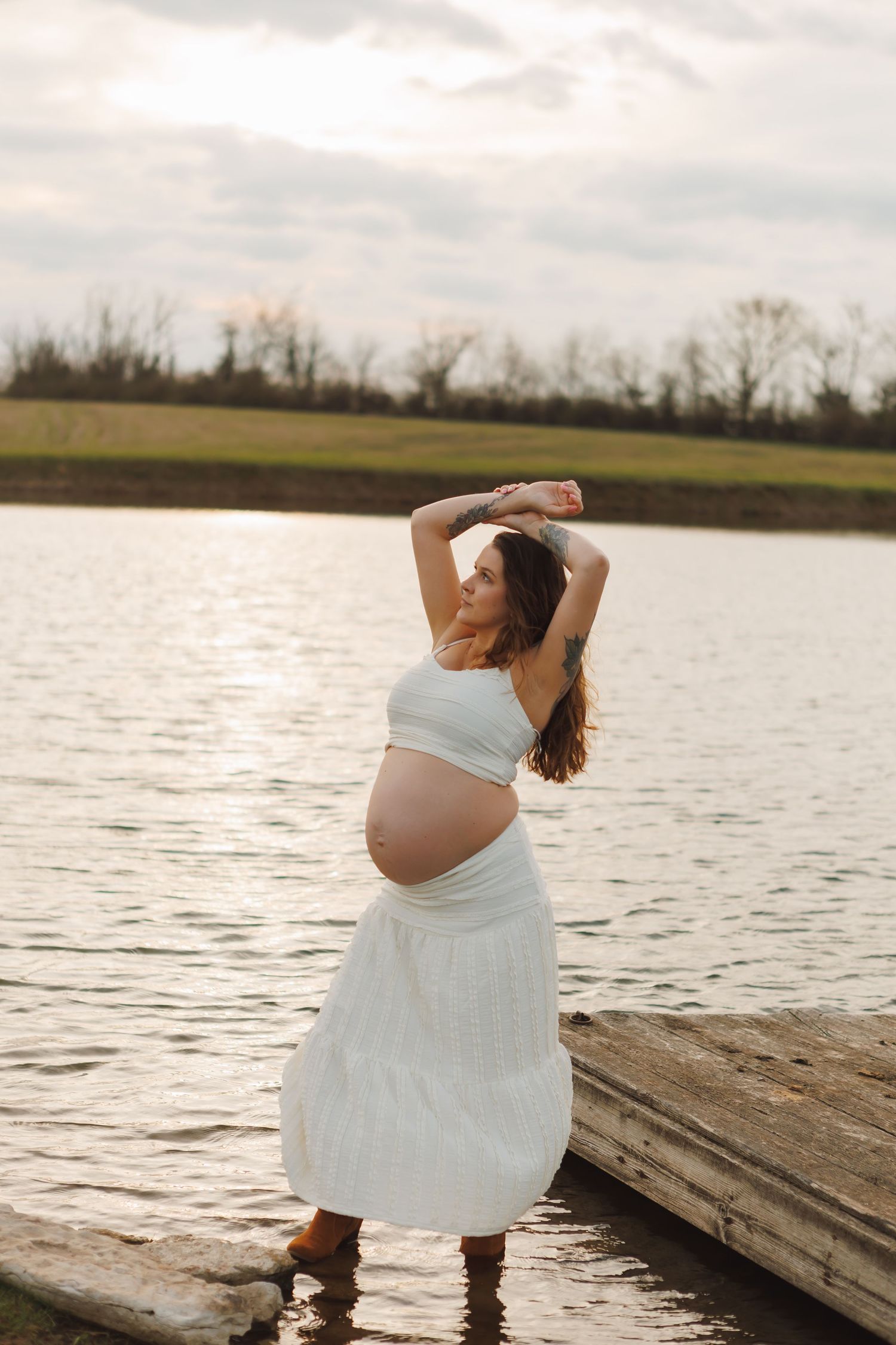 Pregnant woman in white crop top and skirt posing by a lake at sunset with arms raised above her head.