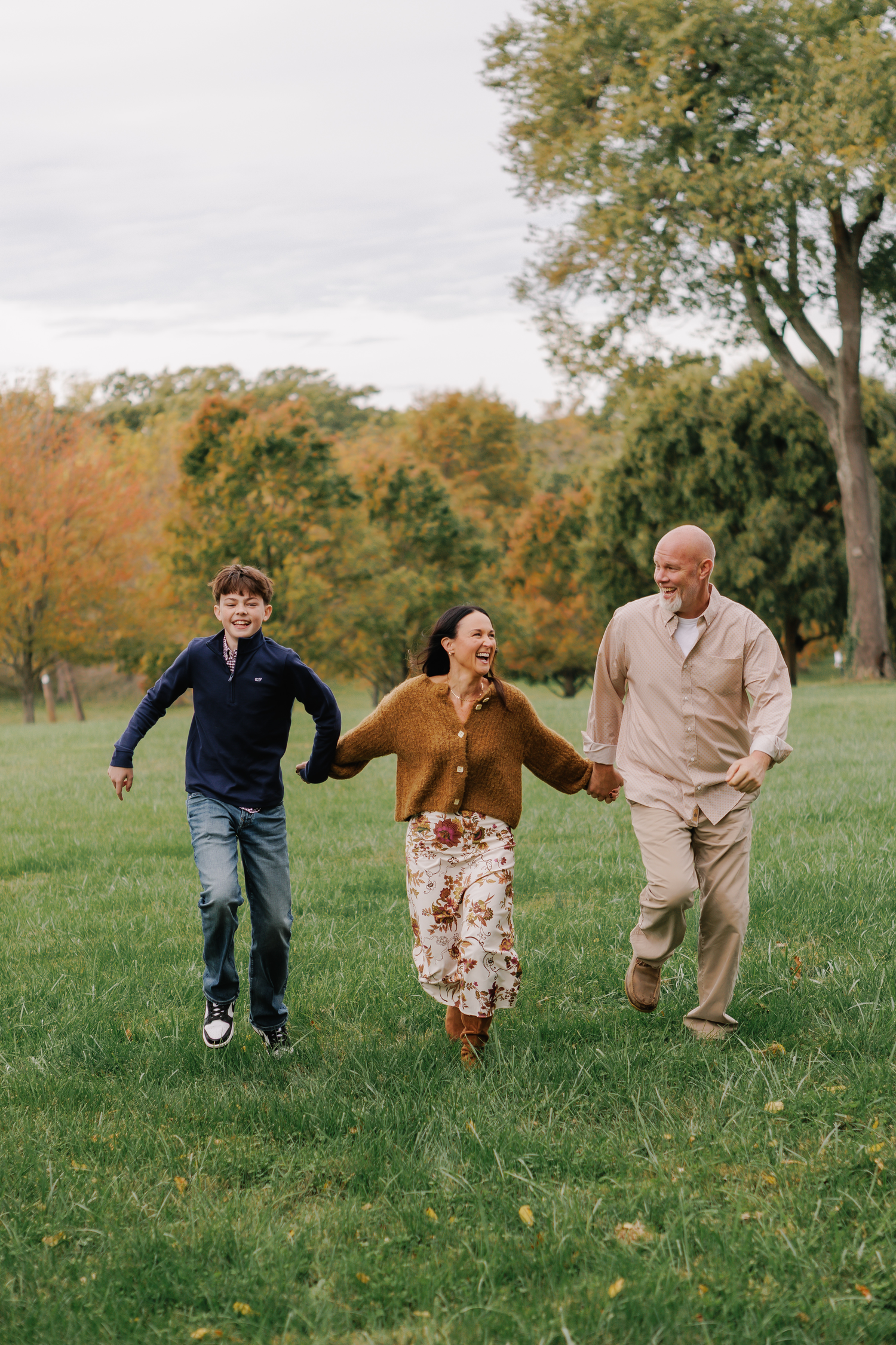 Family of three holding hands and running through a green park with autumn trees in the background.