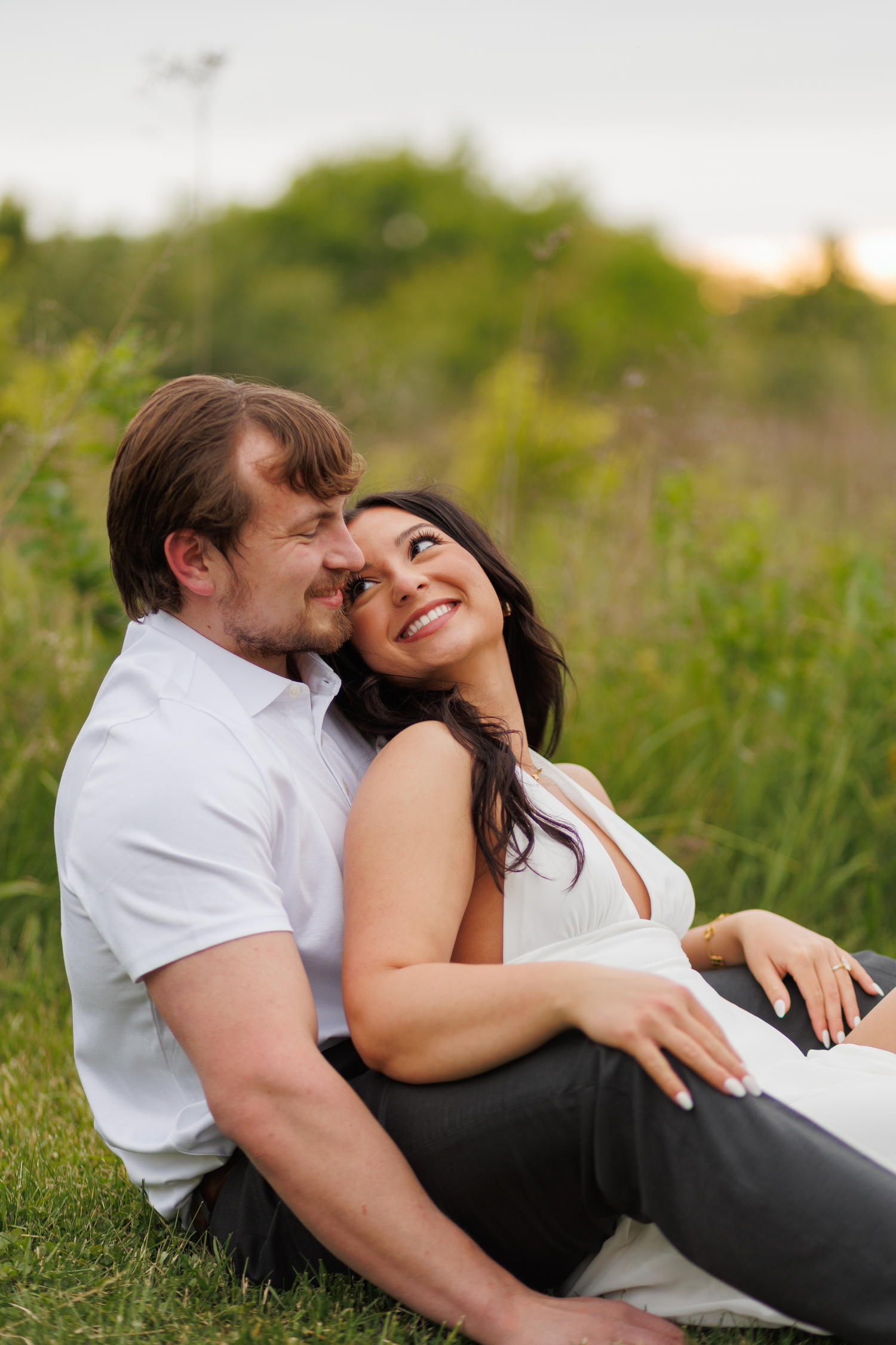 A happy couple sits together in a lush green field, sharing a romantic and joyful moment outdoors.