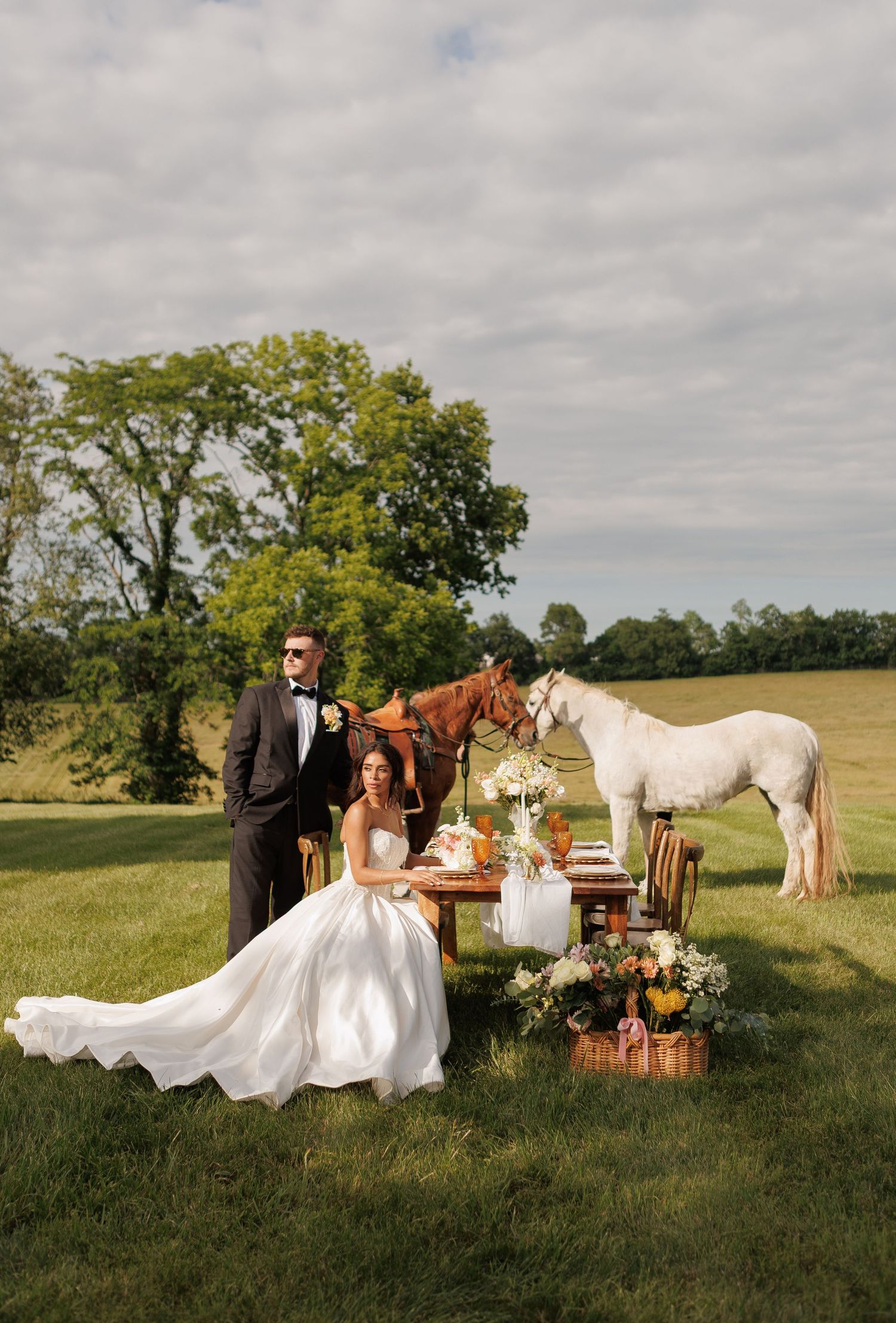 Bride and groom pose at an outdoor wedding table with two horses on a lush green field under a cloudy sky.