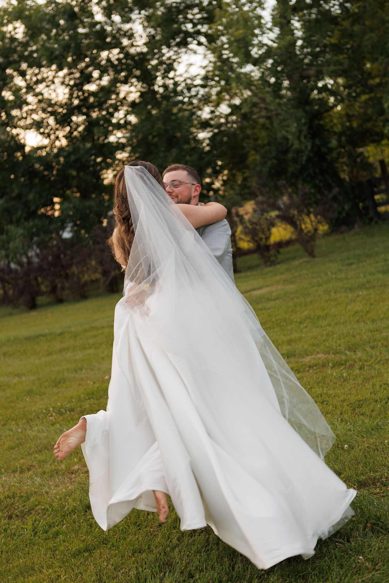A bride in a flowing white gown and veil is lifted and embraced by her groom on a lush green lawn at sunset.