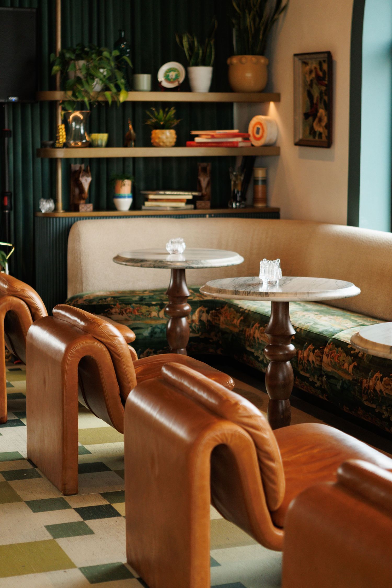 Retro lounge with curved tan leather chairs, marble-top tables, floral sofa, and shelves with plants and decor.