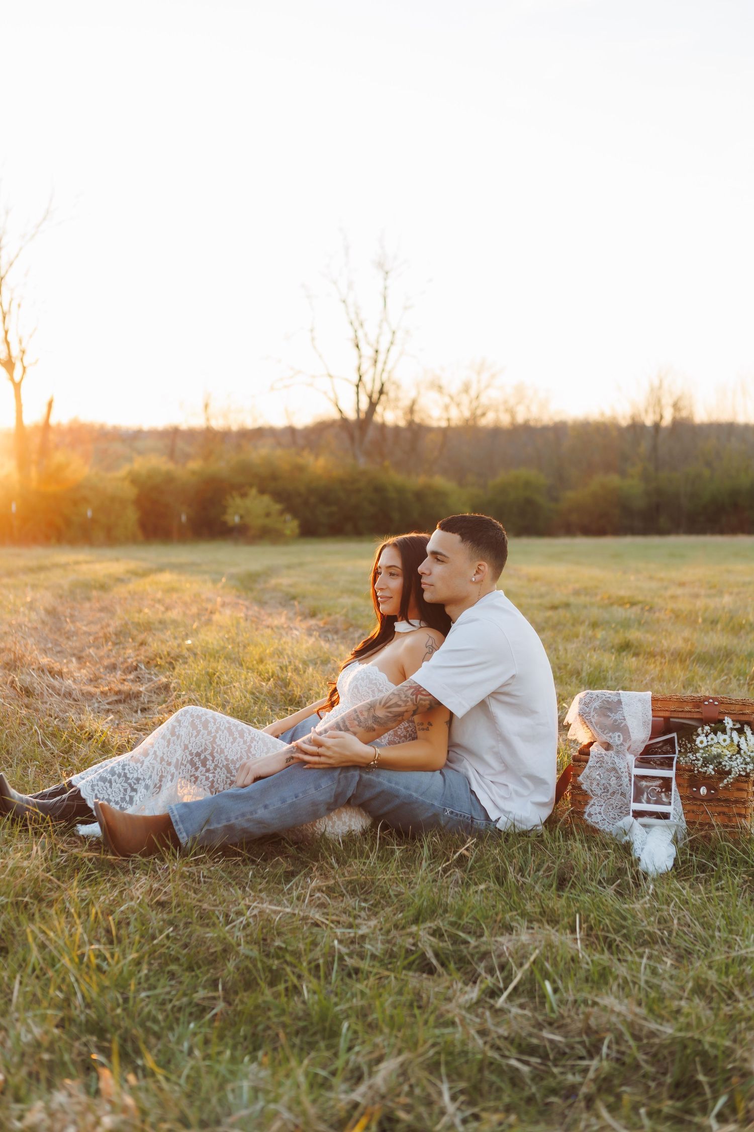Couple sitting together in a golden field at sunset, sharing a tender moment near a picnic basket.
