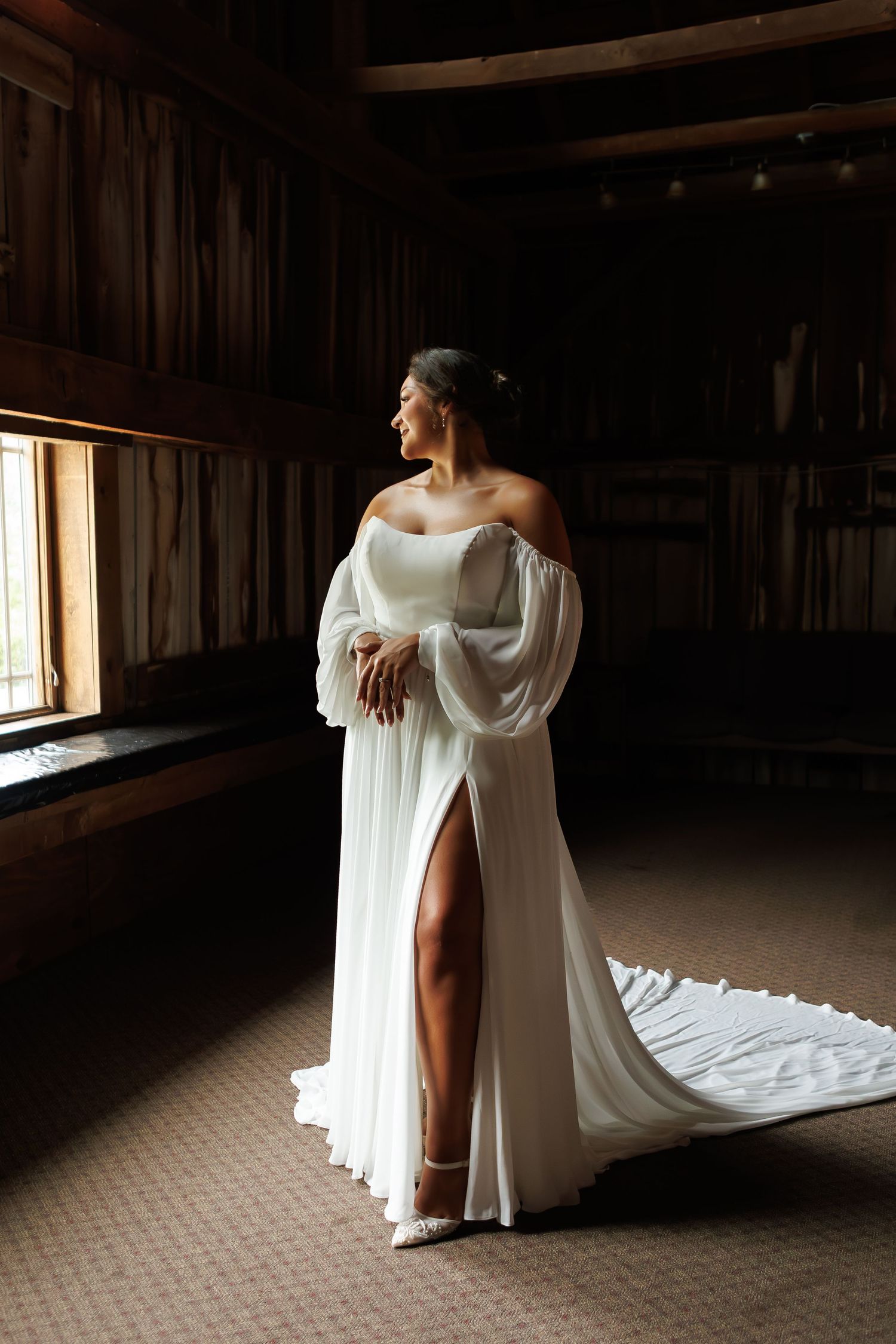 Bride in off-shoulder white gown with flowing sleeves and thigh-high slit stands by window in rustic dark wood venue.