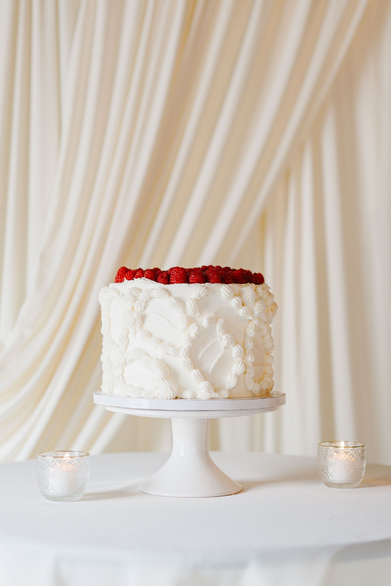 Elegant white cake with red flower topping displayed on pedestal against draped backdrop.