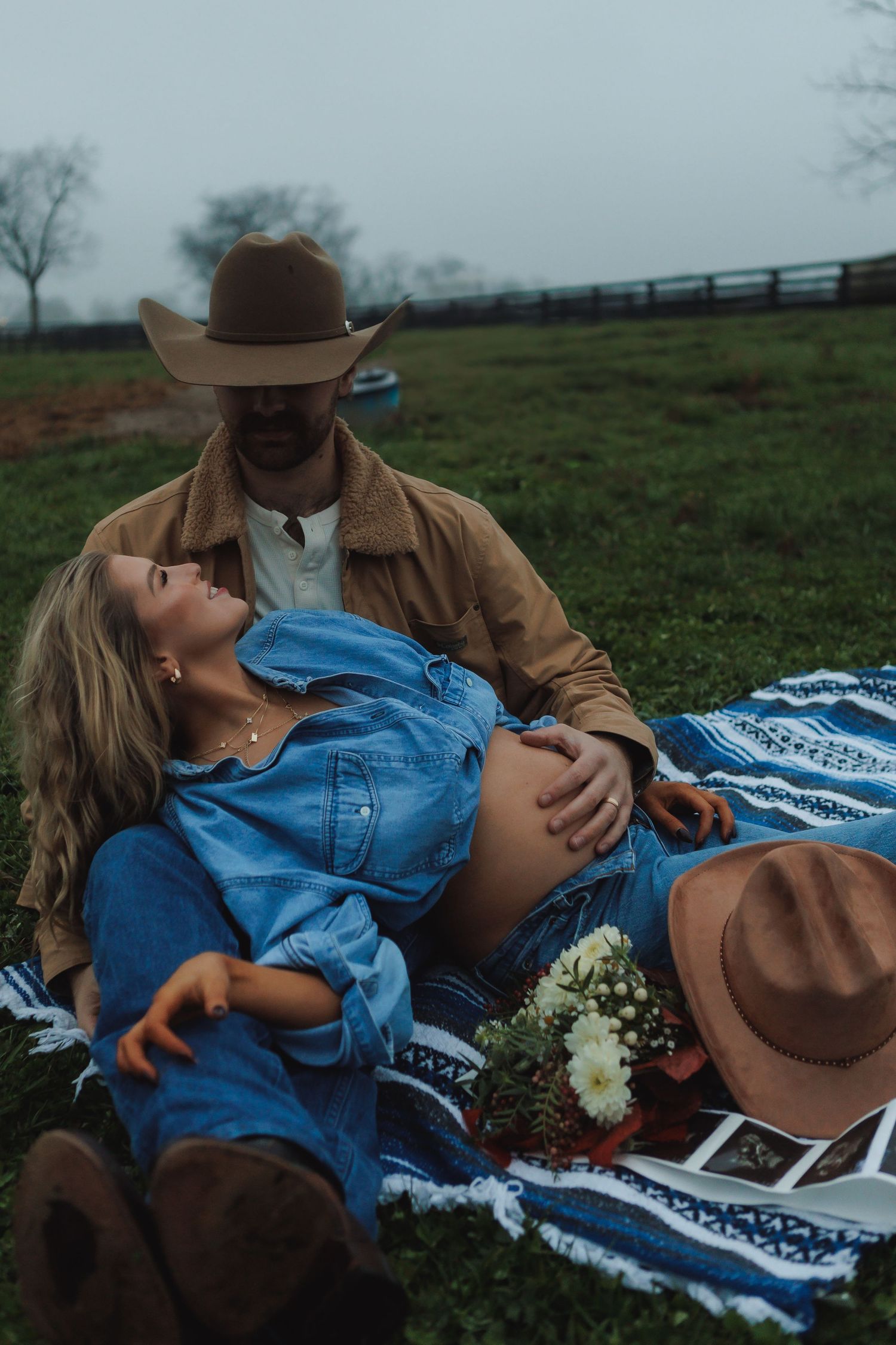 Couple relaxes on blanket with flowers and photos in countryside setting.