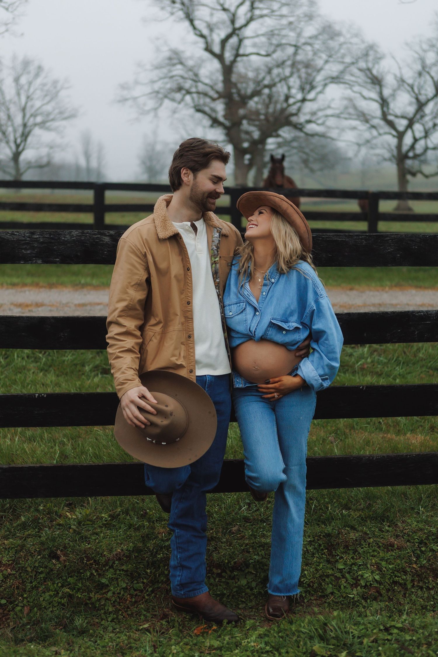 Couple in western wear sharing intimate moment by wooden fence on foggy day.