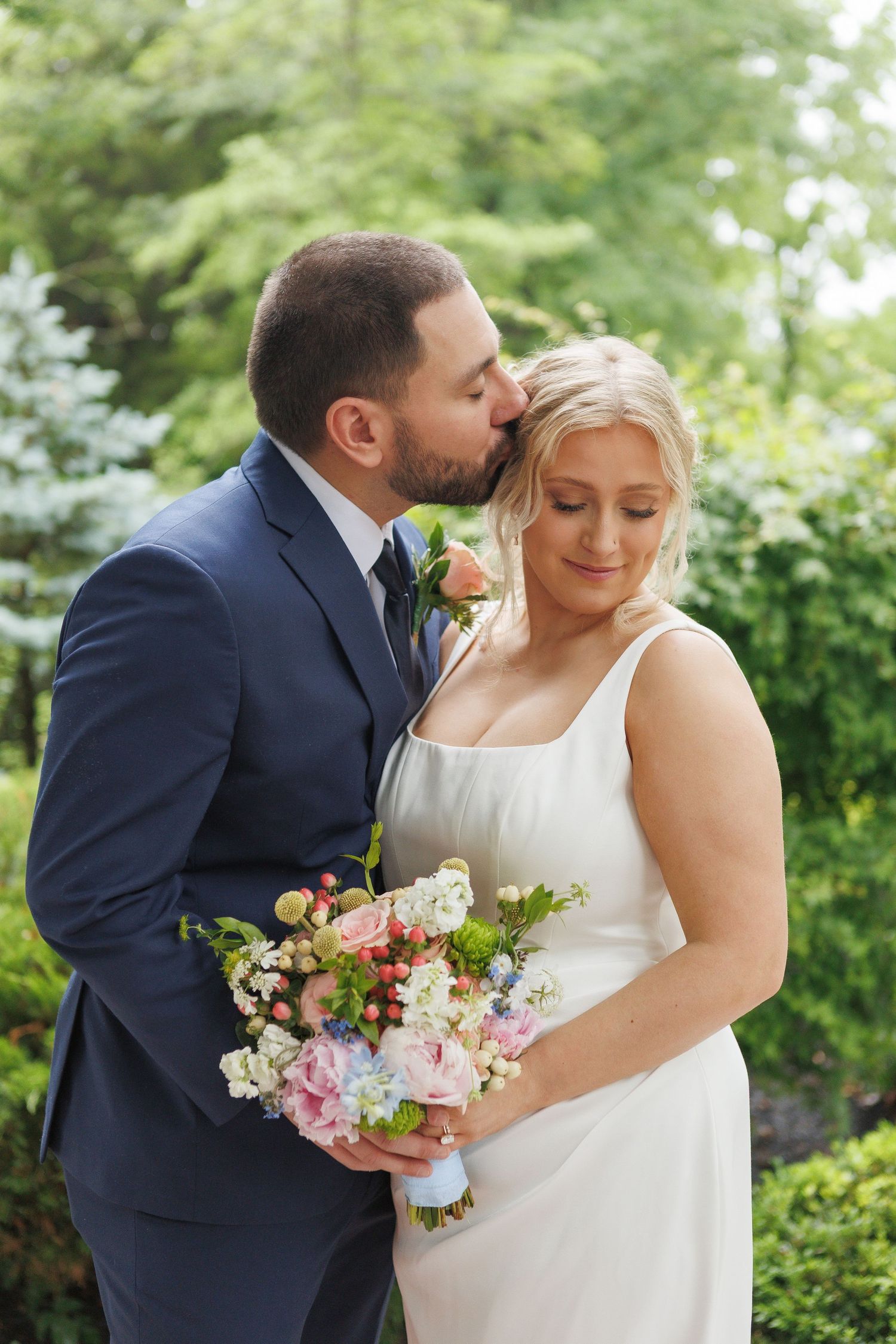 Groom in navy suit kisses bride on forehead as she holds colorful bouquet outdoors amid lush green trees.