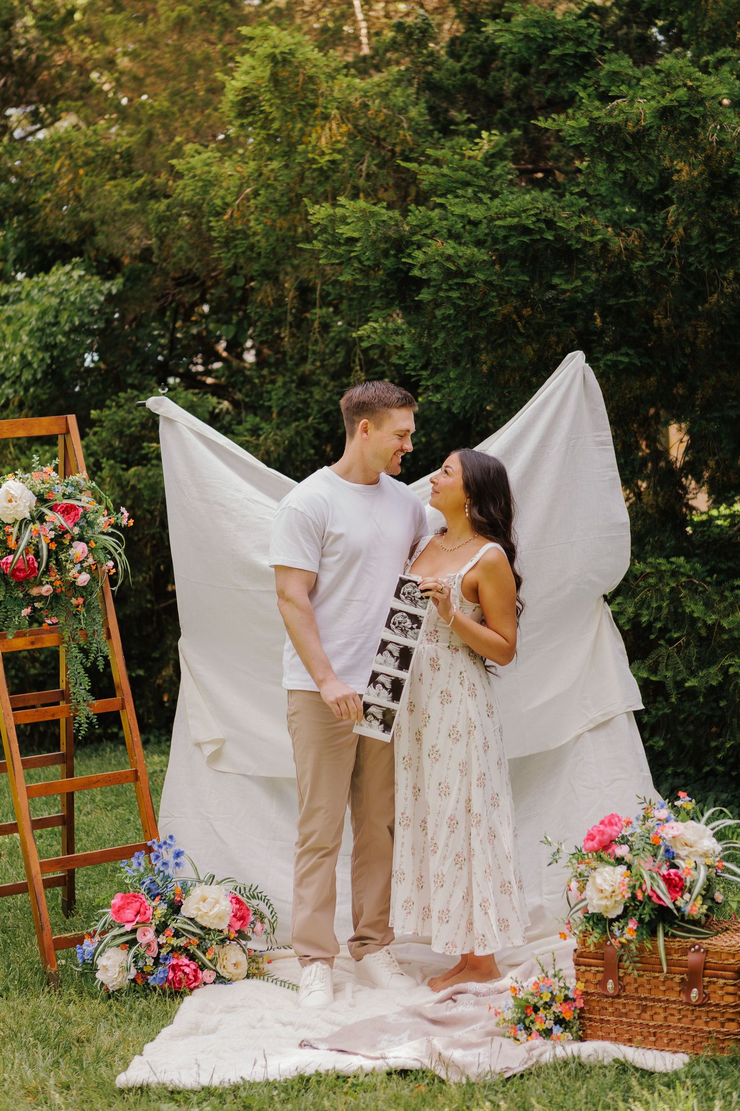 Couple holding ultrasound photos at an outdoor pregnancy announcement with floral decorations and white backdrop.