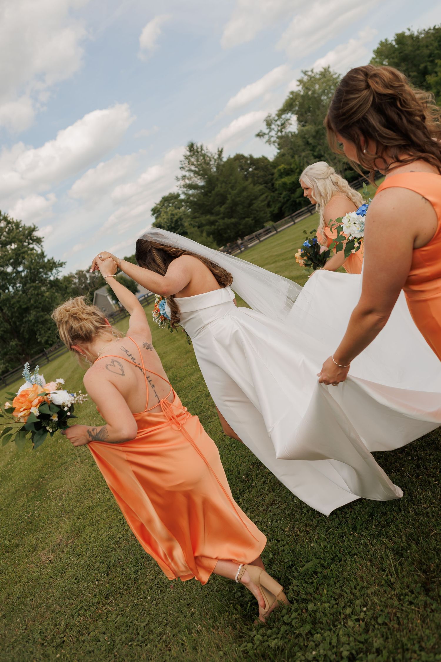 Bridesmaids in orange dresses help a bride in white gown walk across green lawn at outdoor wedding.