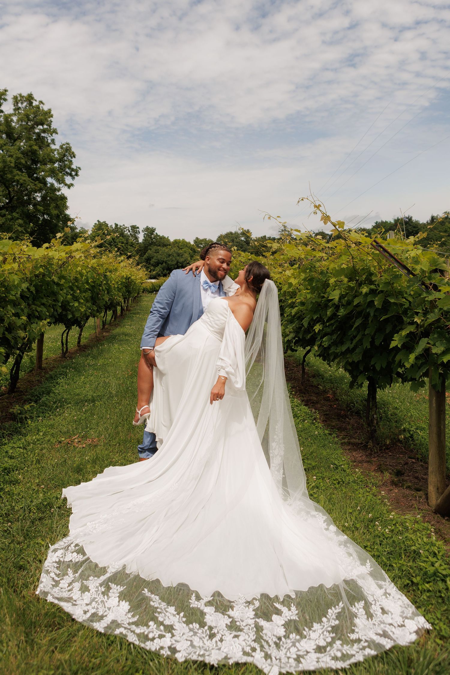 Bride and groom share a romantic kiss in a lush vineyard, her lace cathedral veil trailing along the green grass.