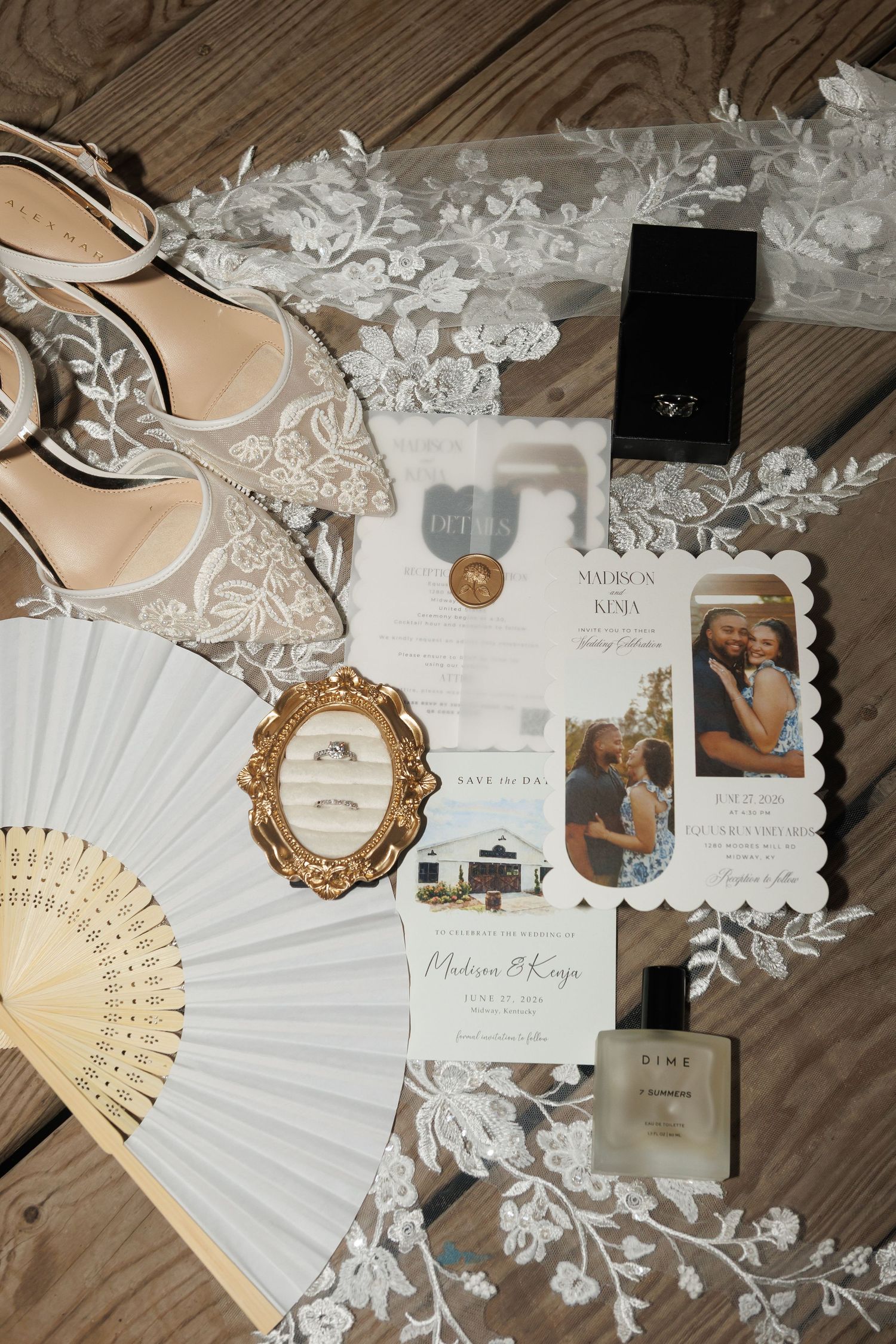 Elegant wedding flat lay with bridal heels, lace veil, rings, invitations, perfume, and hand fan on wood.