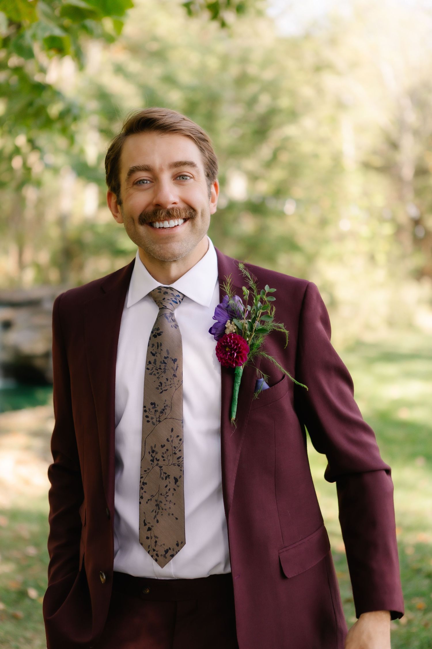 Men wearing burgundy suits and patterned gray ties adorned with matching floral boutonnieres in an outdoor setting.