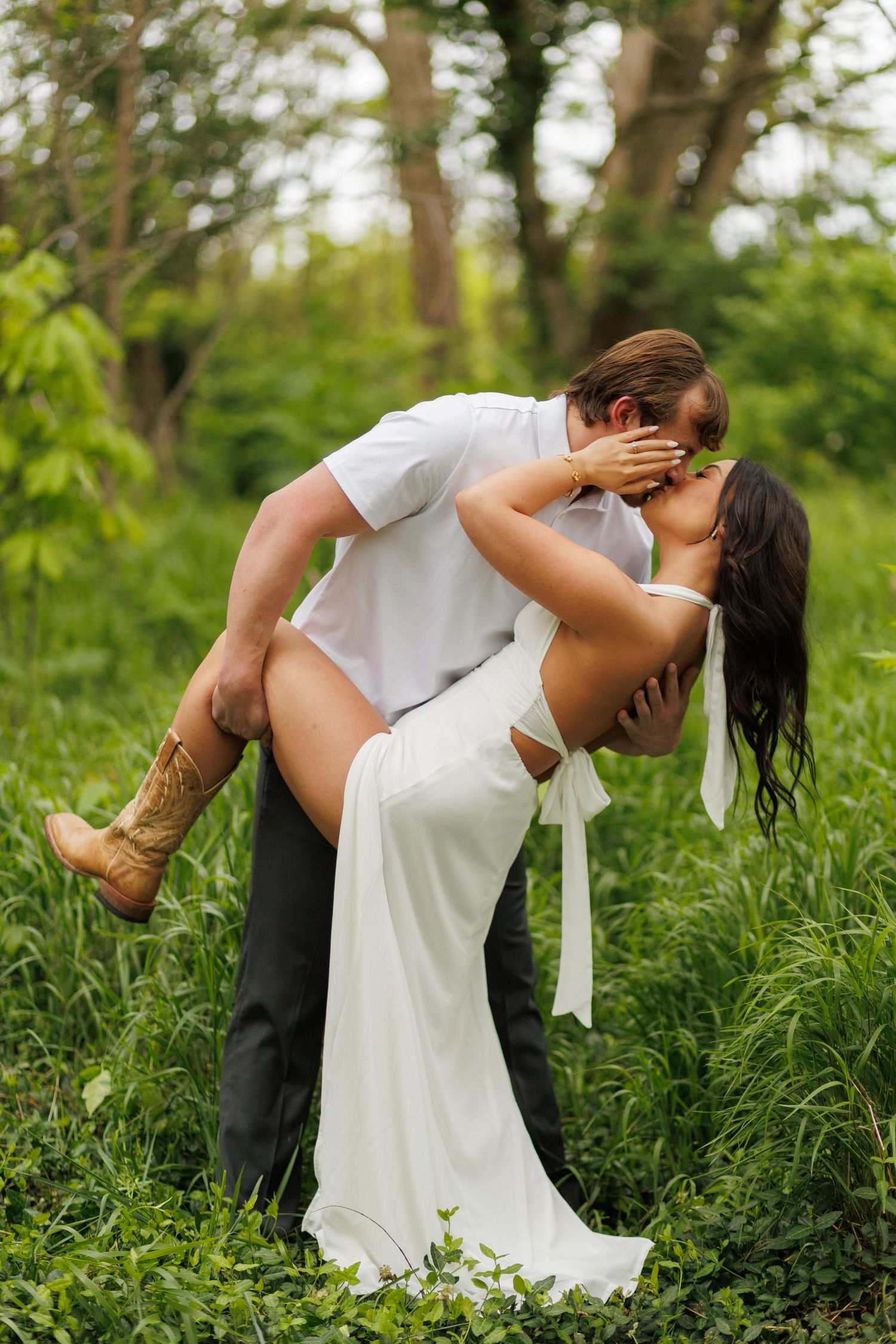 A couple shares a romantic kiss outdoors, the bride in a white dress and boots, groom in white shirt, surrounded by lush greenery.