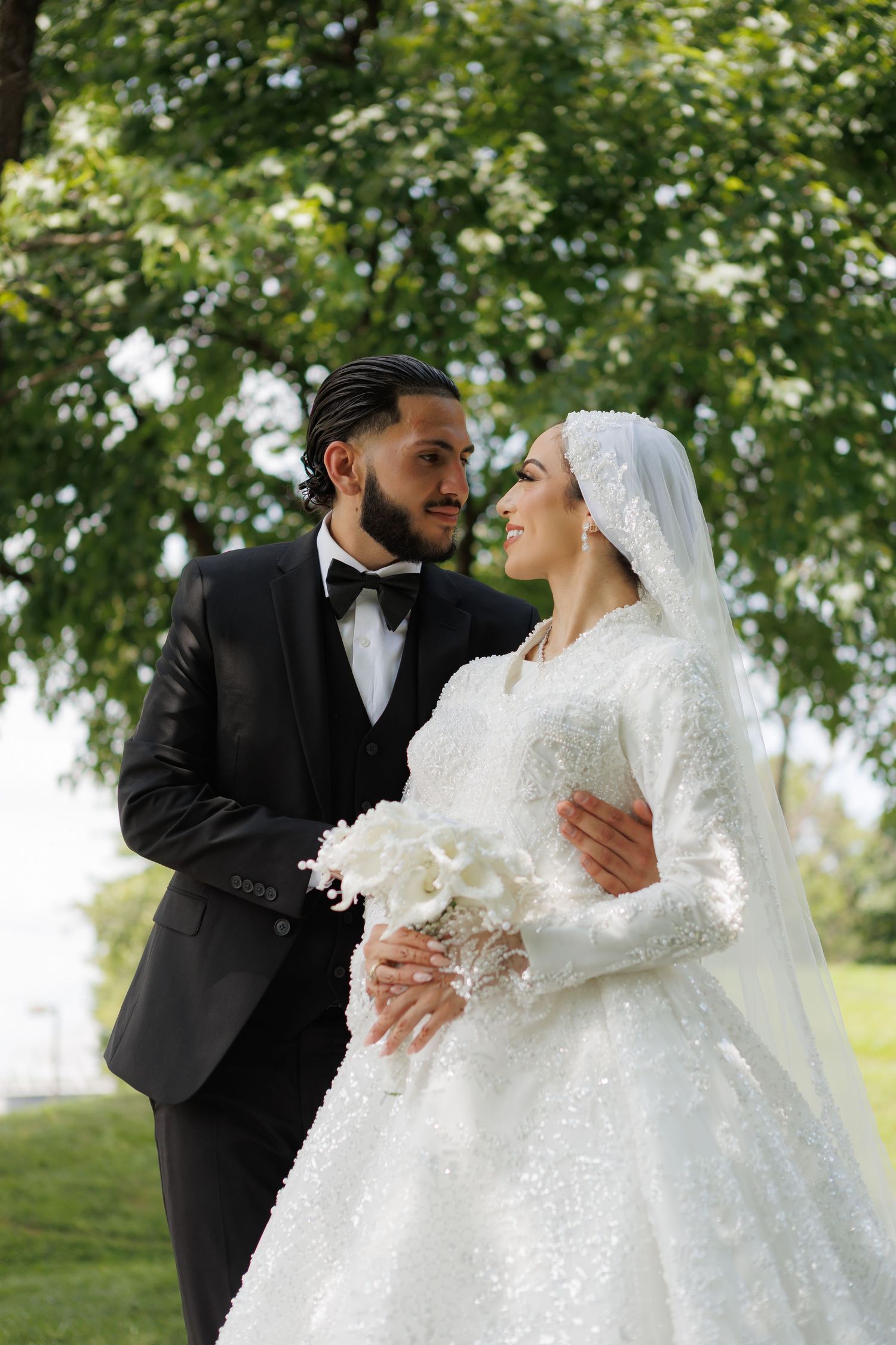A bride in white lace gown and veil holds flowers as groom in black tuxedo embraces her outdoors.