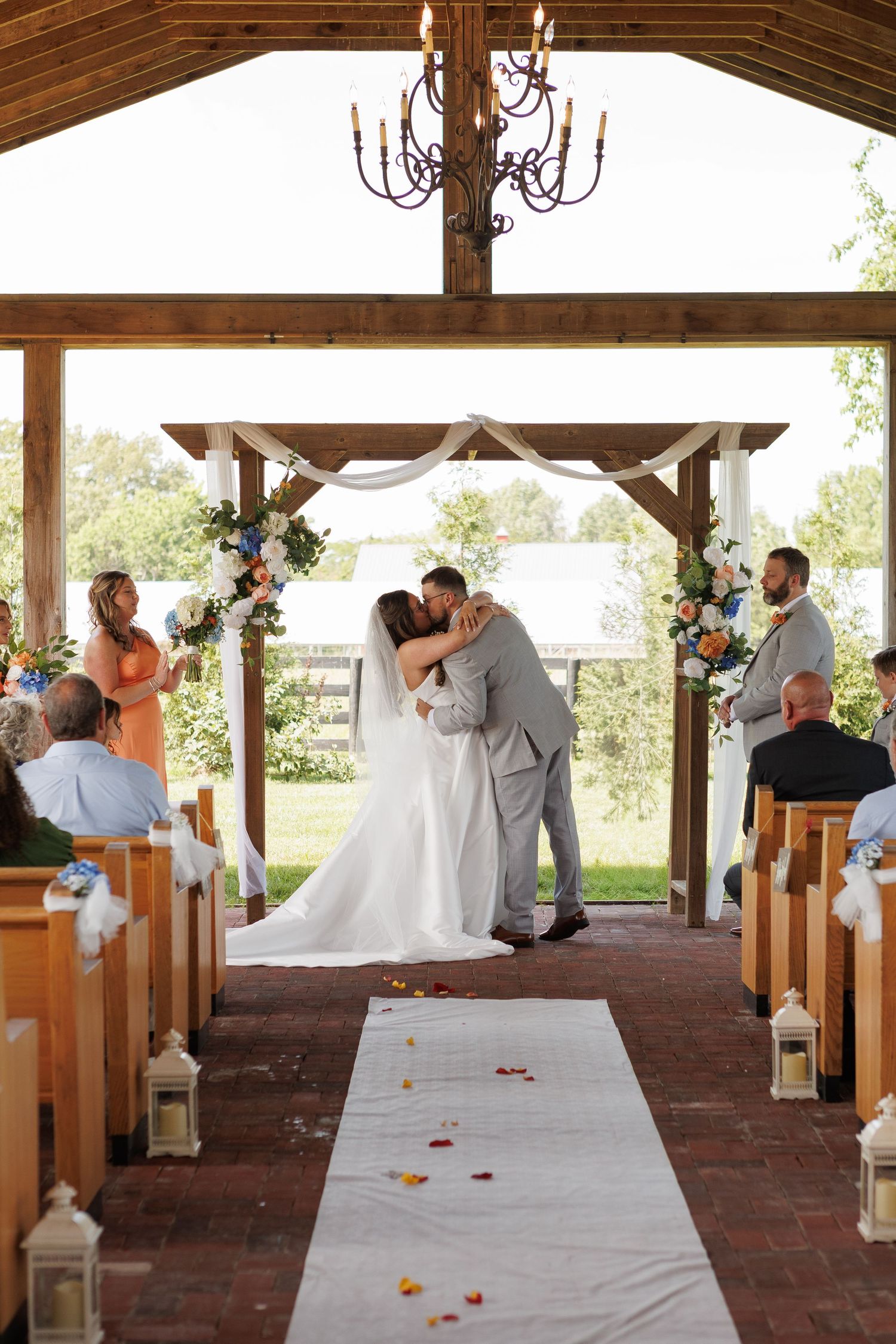 Bride and groom share first kiss at outdoor chapel wedding ceremony with floral arch, chandelier, and rose petal aisle.