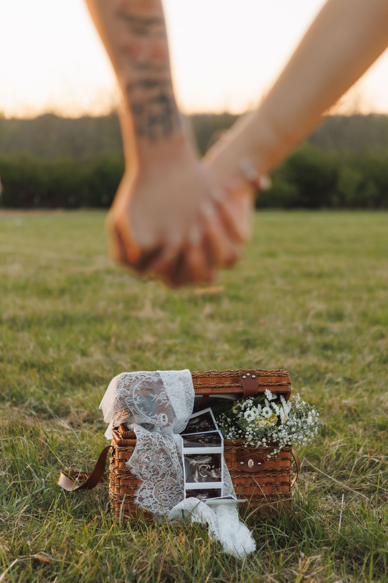 Couple holding hands over a wicker basket with lace, flowers, and photos on grass at golden hour.