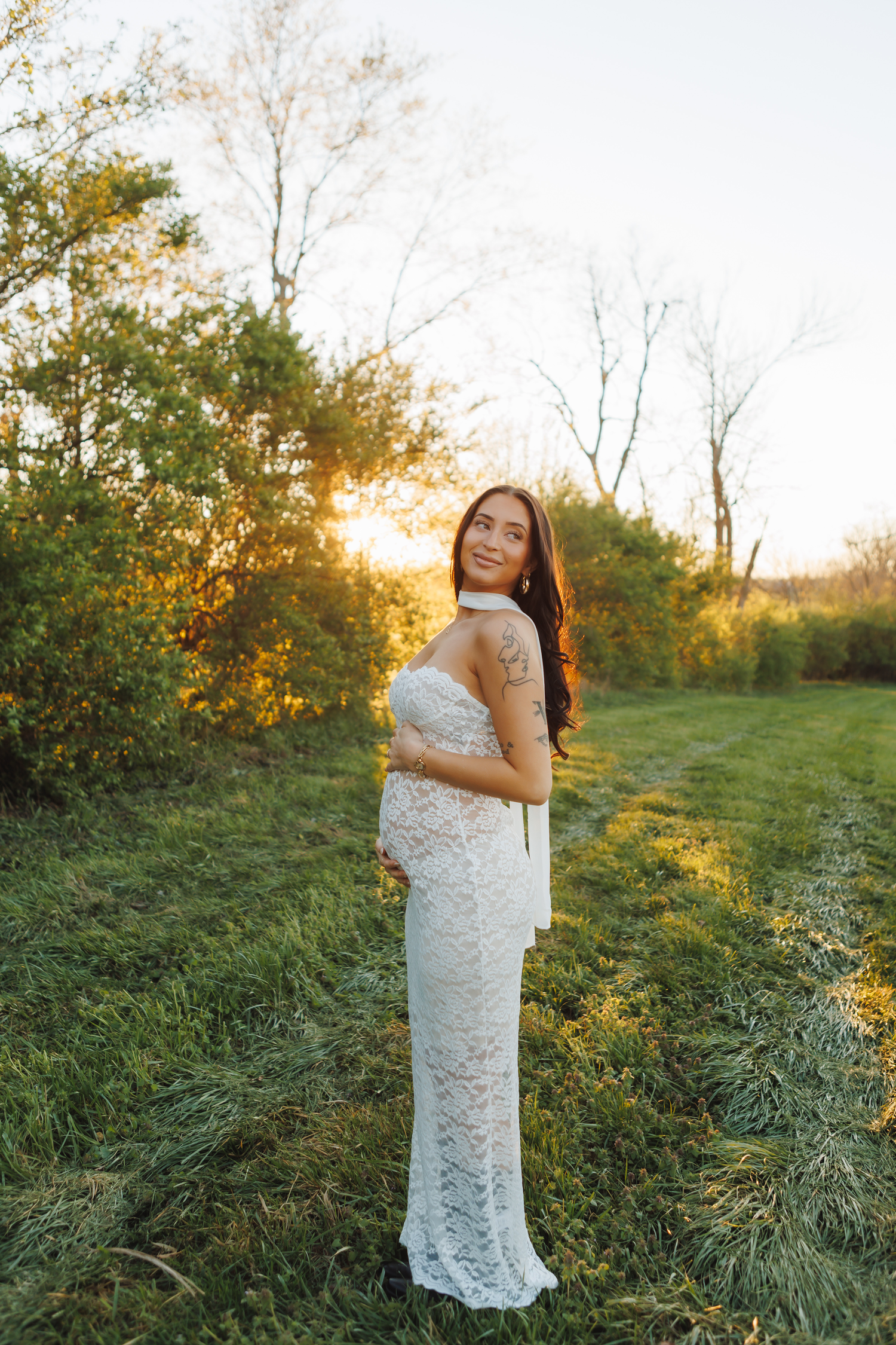 Pregnant woman in white lace dress standing in green field at golden sunset hour.