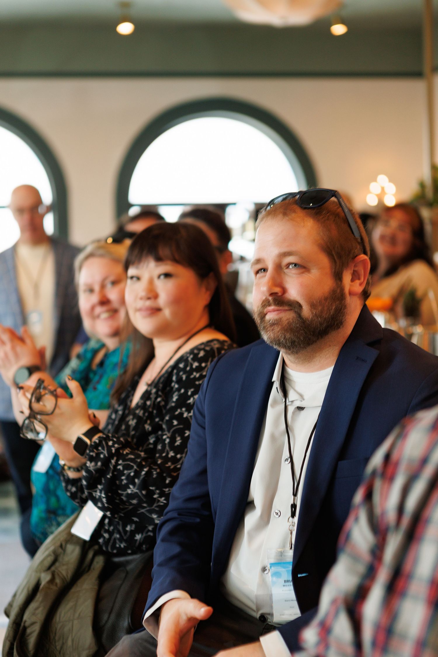 Audience members seated at an indoor event, including a bearded man in a navy blazer with a lanyard and sunglasses on his head.