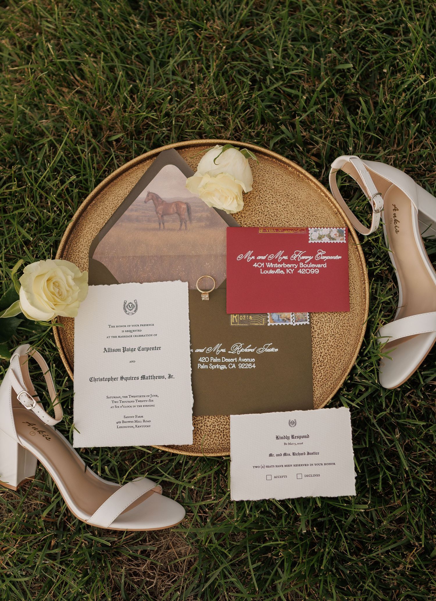 Elegant wedding invitation suite with horse motif, white heels, and cream roses on a gold tray in green grass.