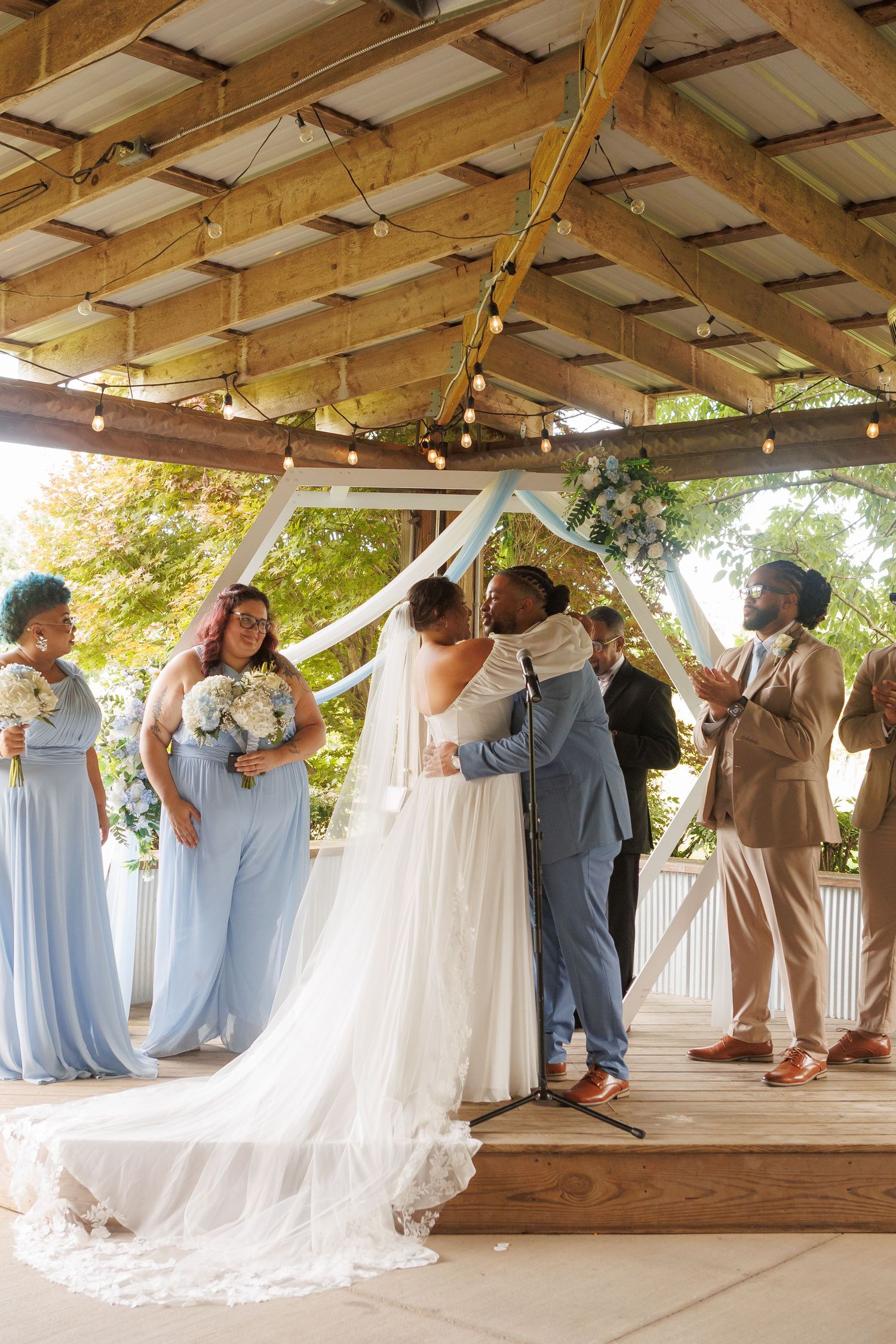 Bride and groom share first kiss under floral arch, flanked by bridesmaids in blue and groomsmen in tan suits.