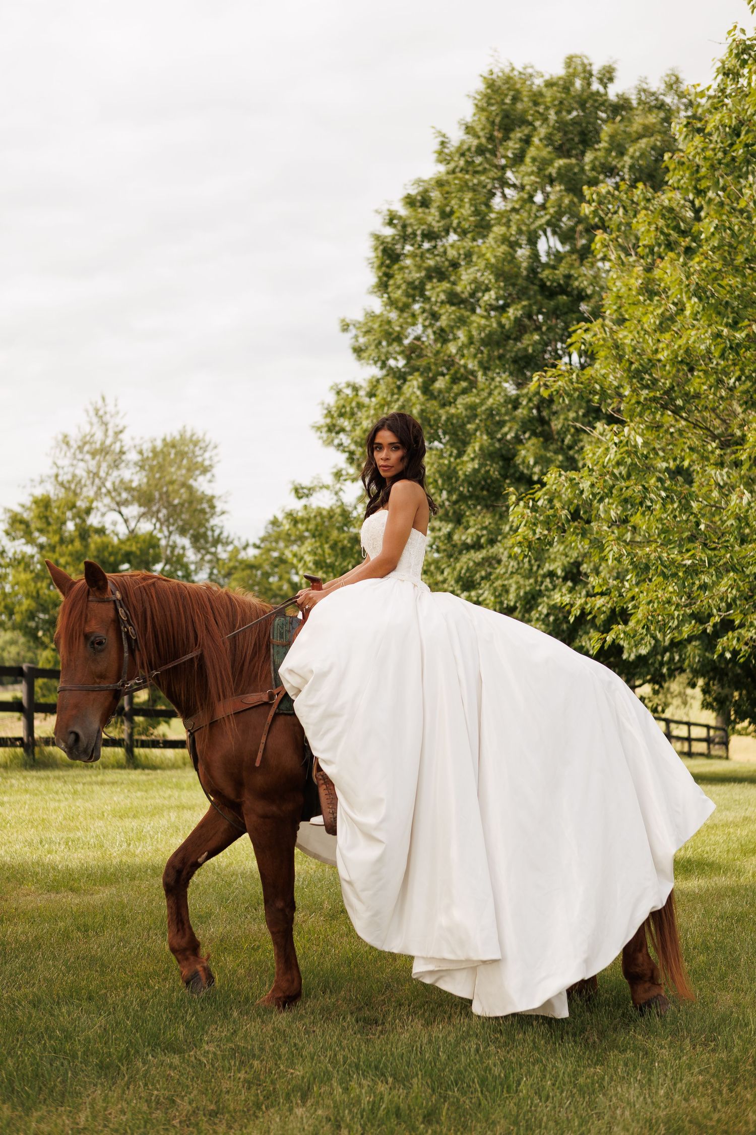 Elegant bride in strapless white ball gown sits atop a chestnut horse on a green farm.