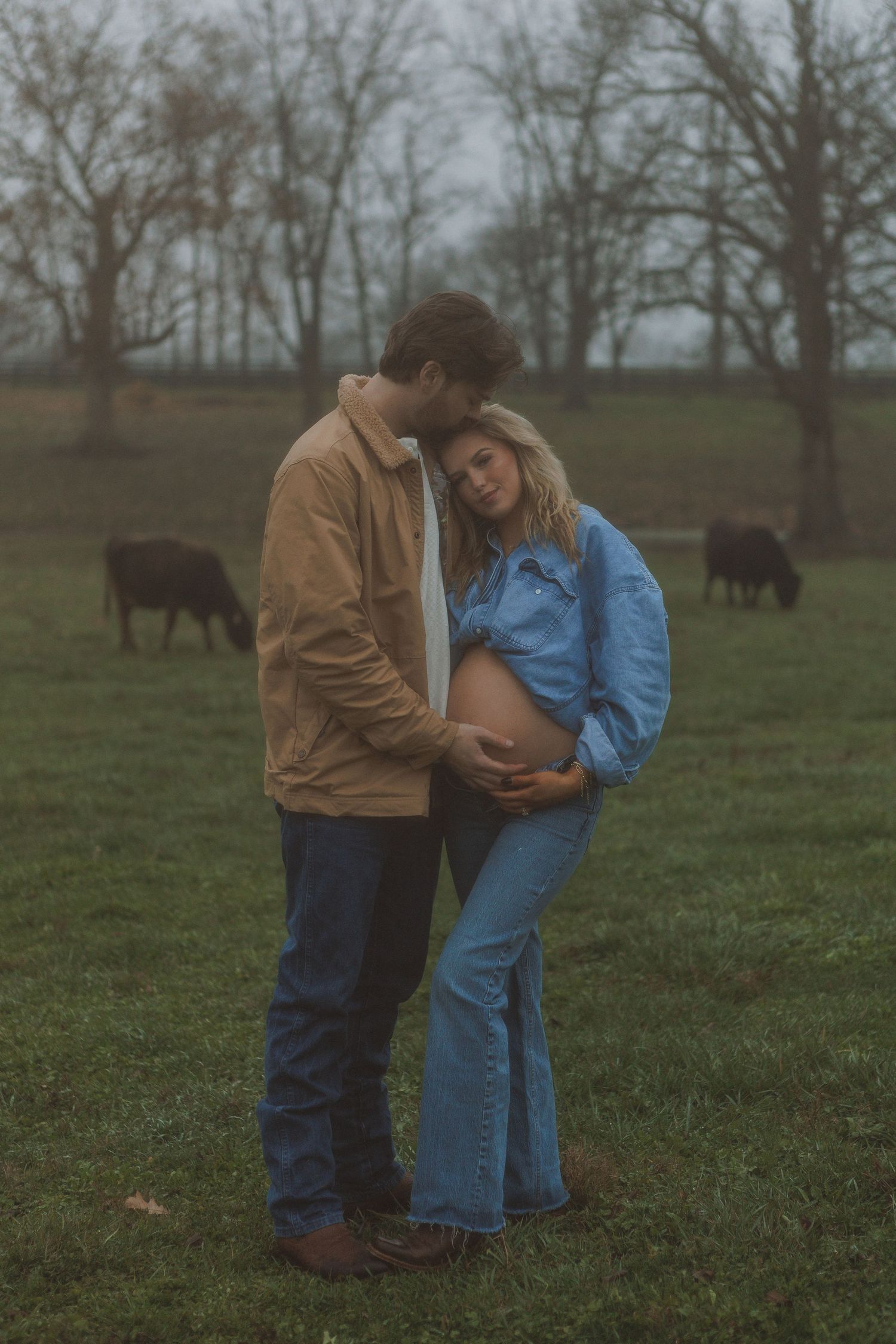 A couple in denim and brown attire share an intimate moment in a rural field with cattle in the background.