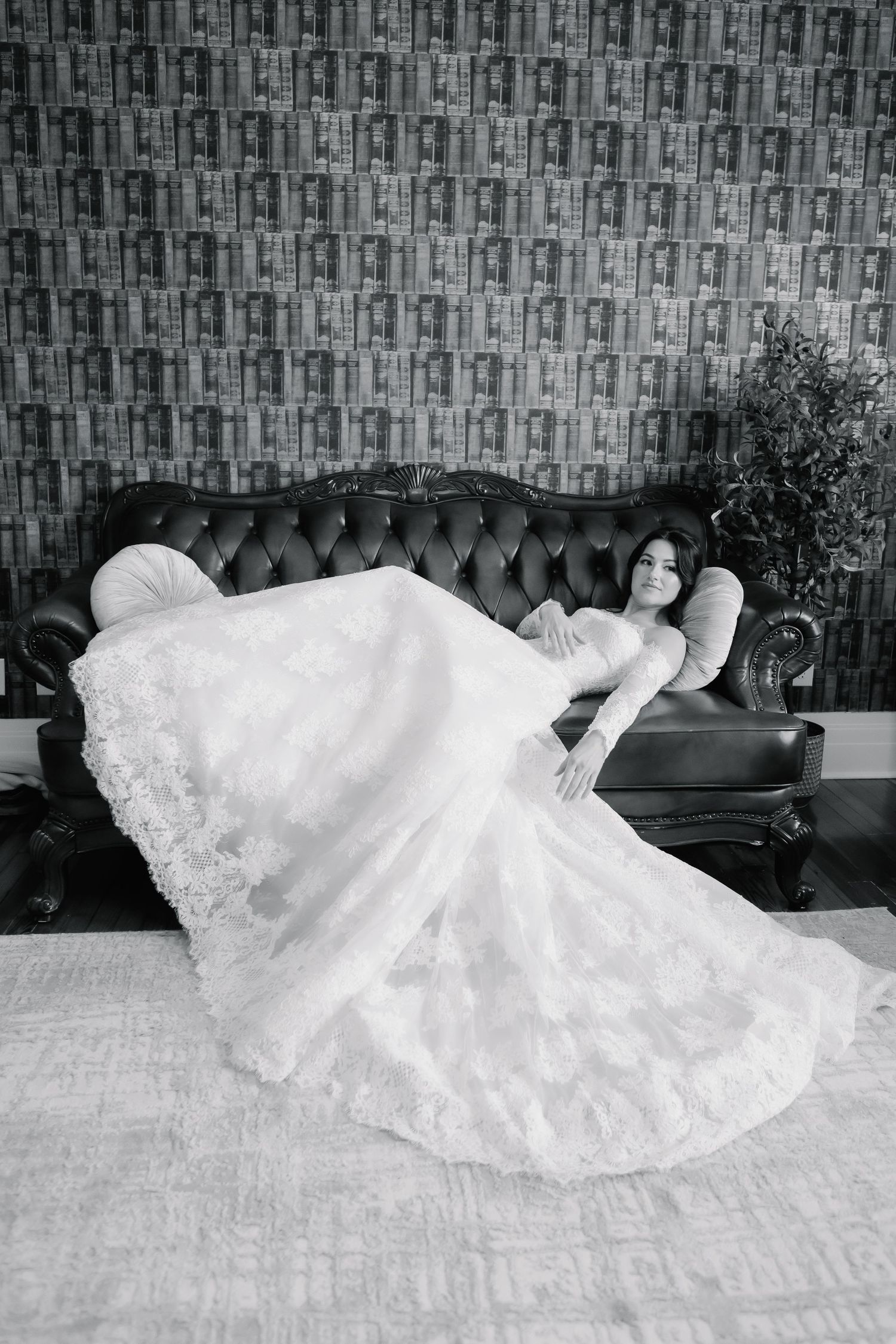 Elegant bride in a white lace wedding gown reclining on a tufted leather sofa in a black and white portrait.