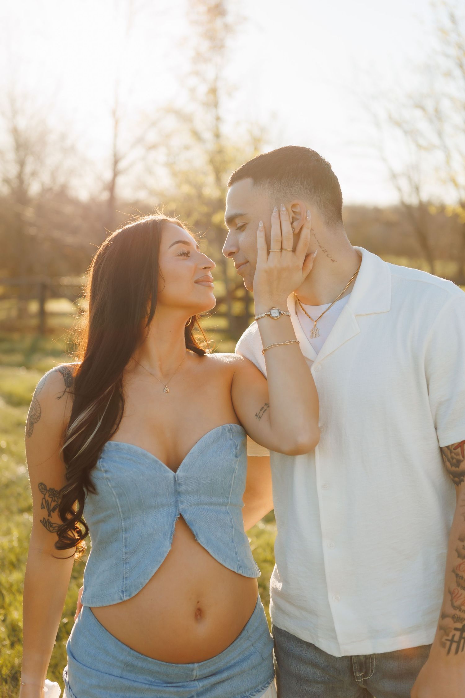 Couple sharing a tender moment outdoors at golden hour, woman in denim crop top with tattoos, man in white shirt.