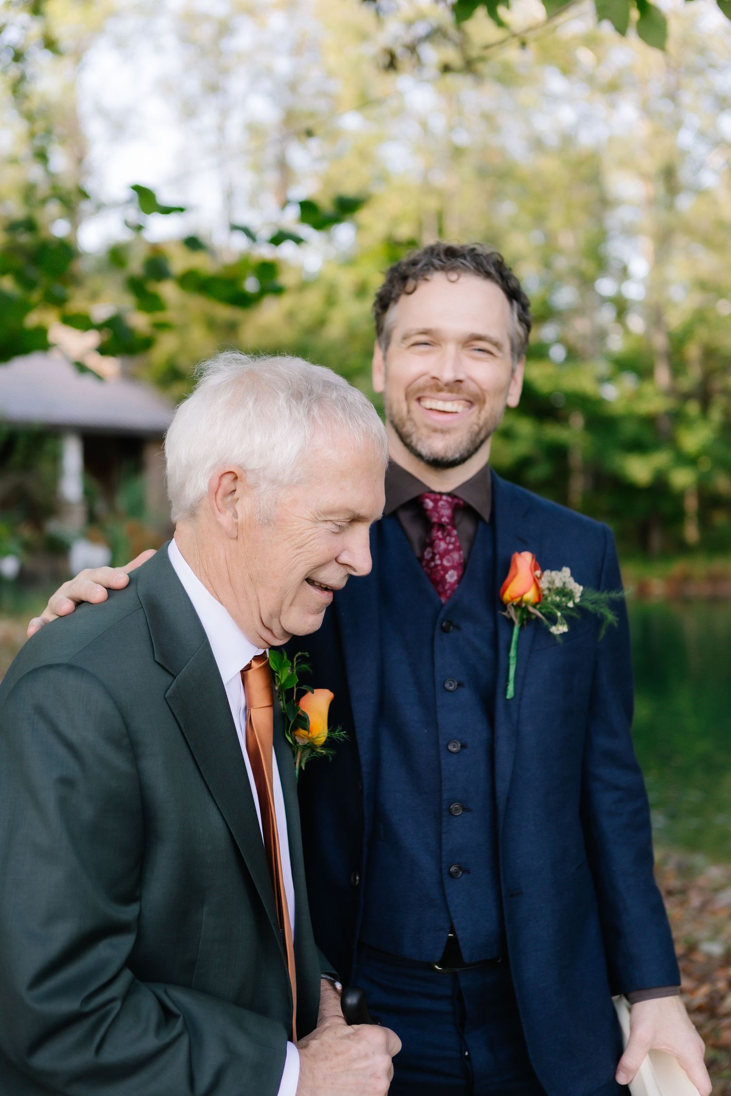 Candid outdoor moment between wedding party members wearing dark suits and boutonnieres.
