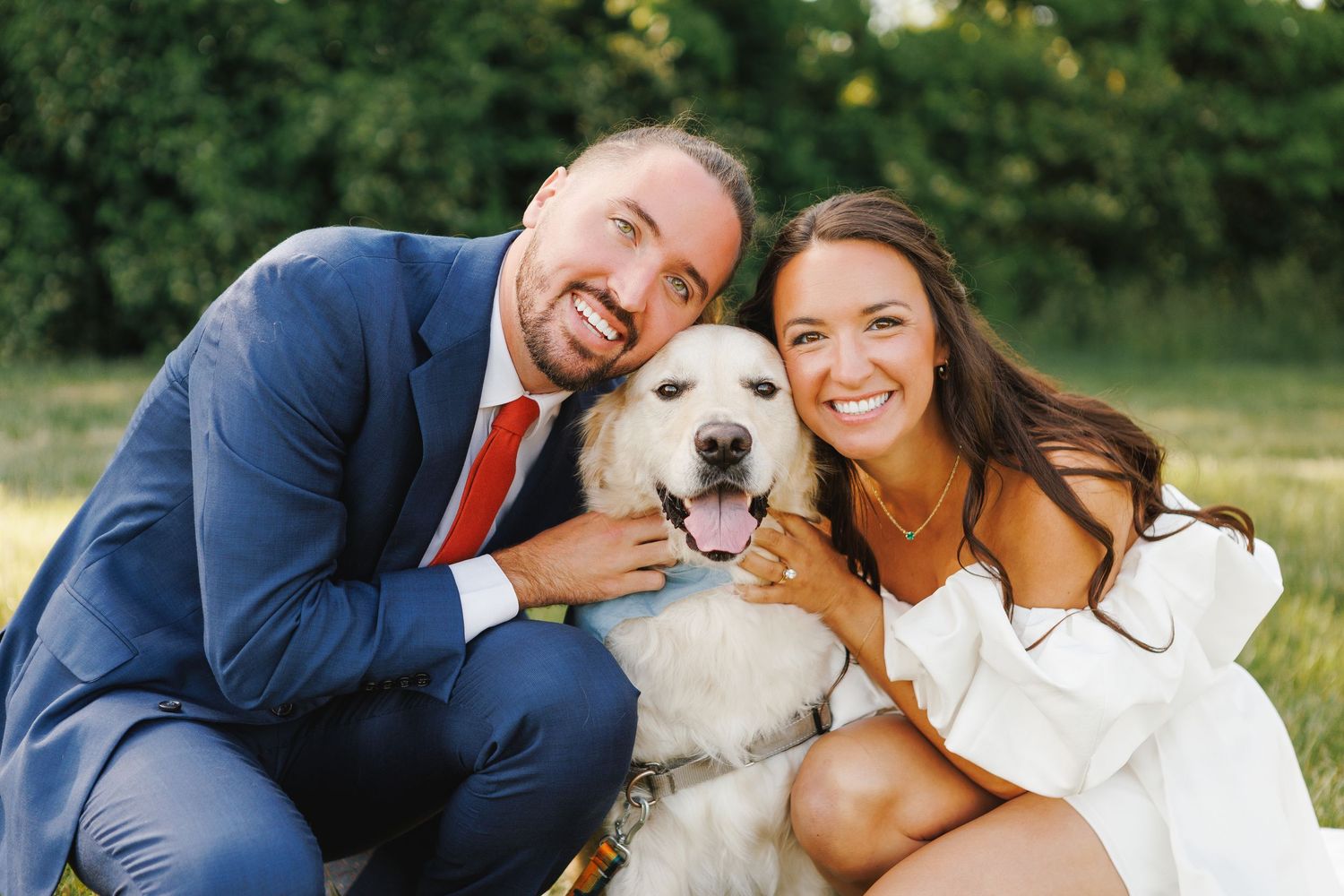 Smiling couple in formal attire posing outdoors with their fluffy white golden retriever dog.