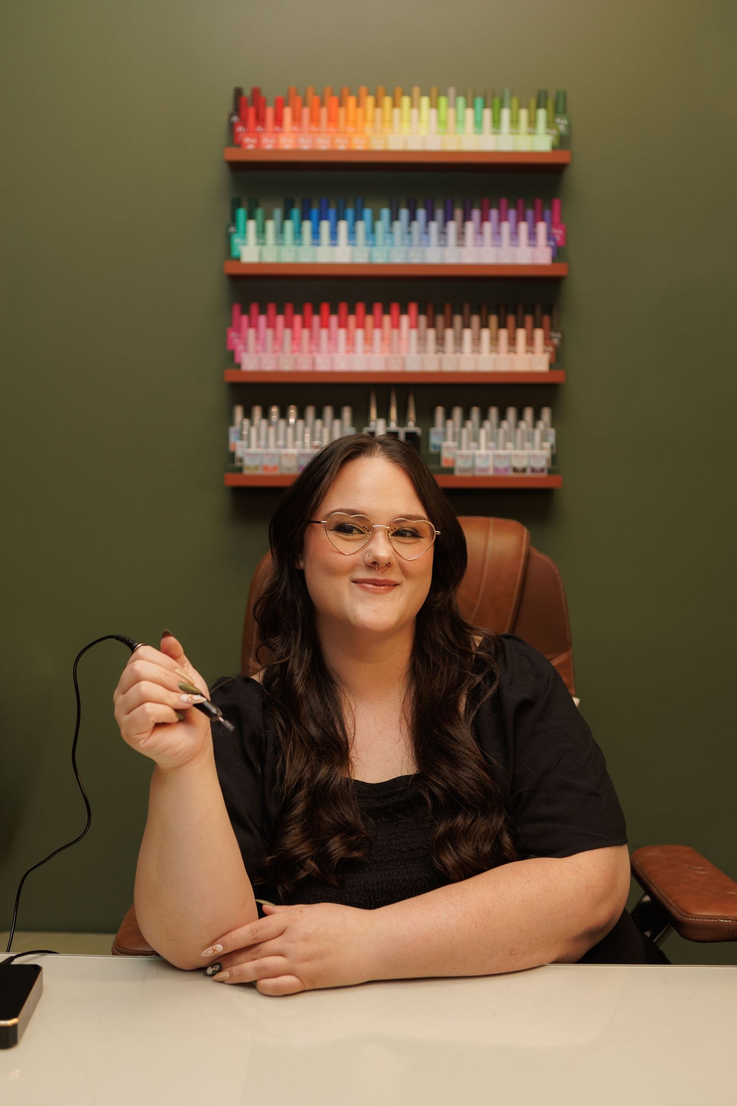 Nail technician sitting at salon desk holding nail tool, with colorful nail polish display on green wall behind her.
