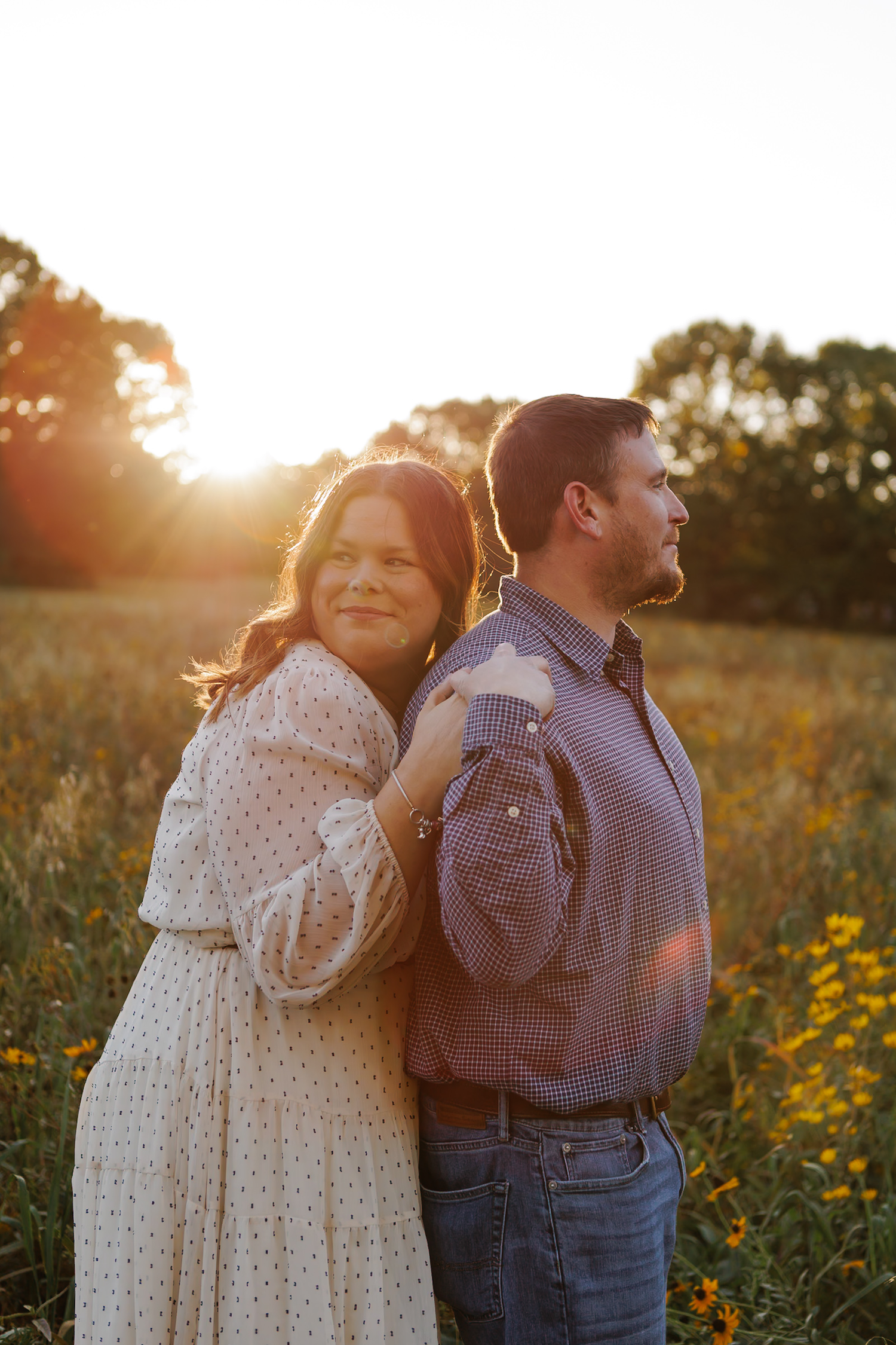 Couple poses back-to-back in golden field at sunset, woman in floral dress smiling, man in plaid shirt with suspenders.
