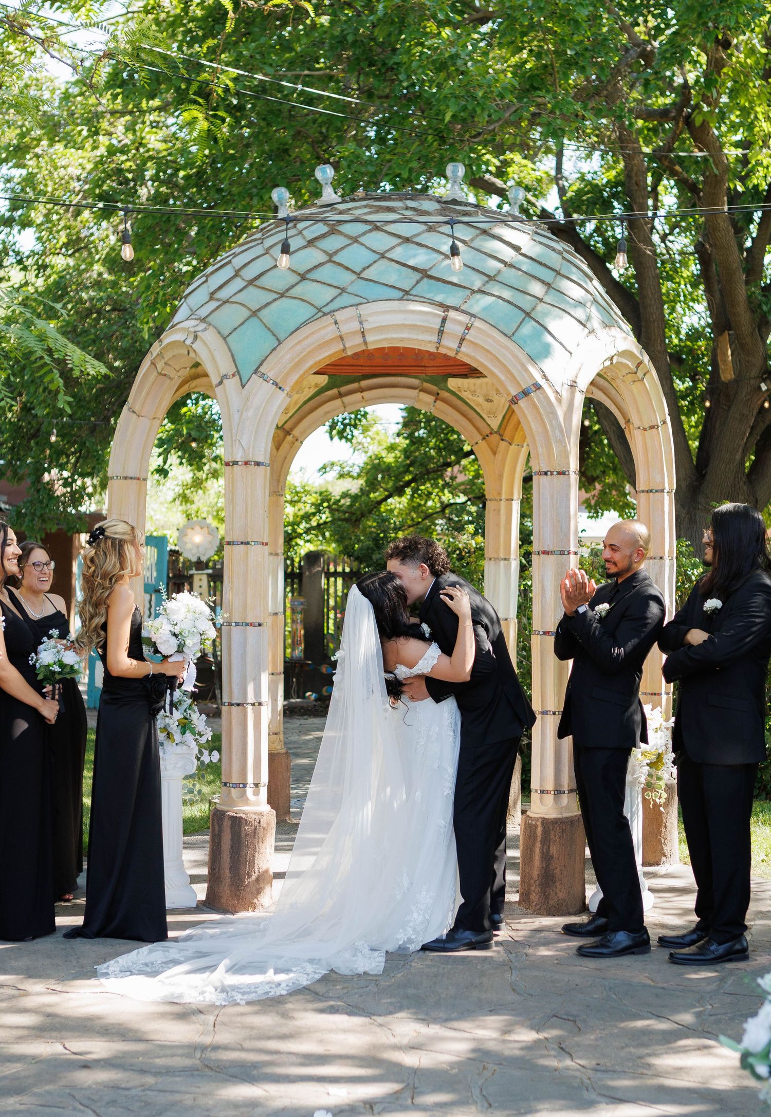 Bride and groom share first kiss under elegant arch gazebo outdoors, flanked by bridesmaids in black dresses.
