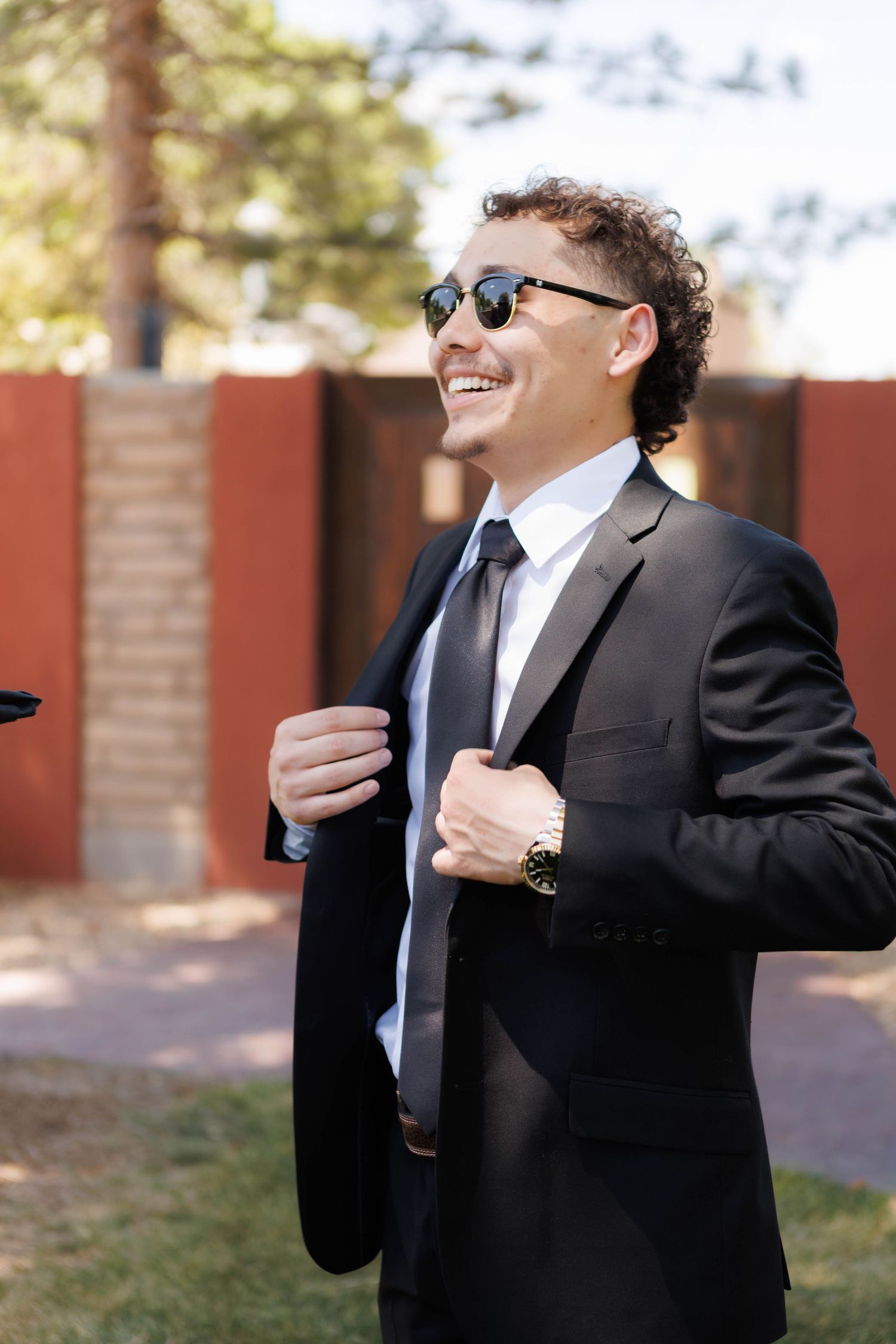 Smiling man in black suit, tie, and sunglasses adjusting jacket outdoors on sunny day.
