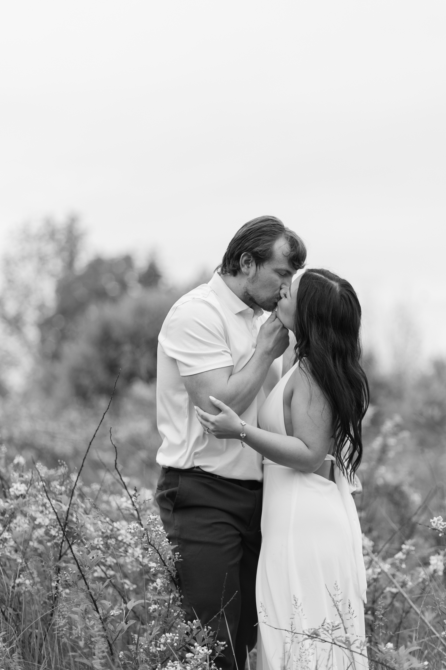 Black and white photo of a romantic couple almost kissing in a wildflower field, woman in white dress.