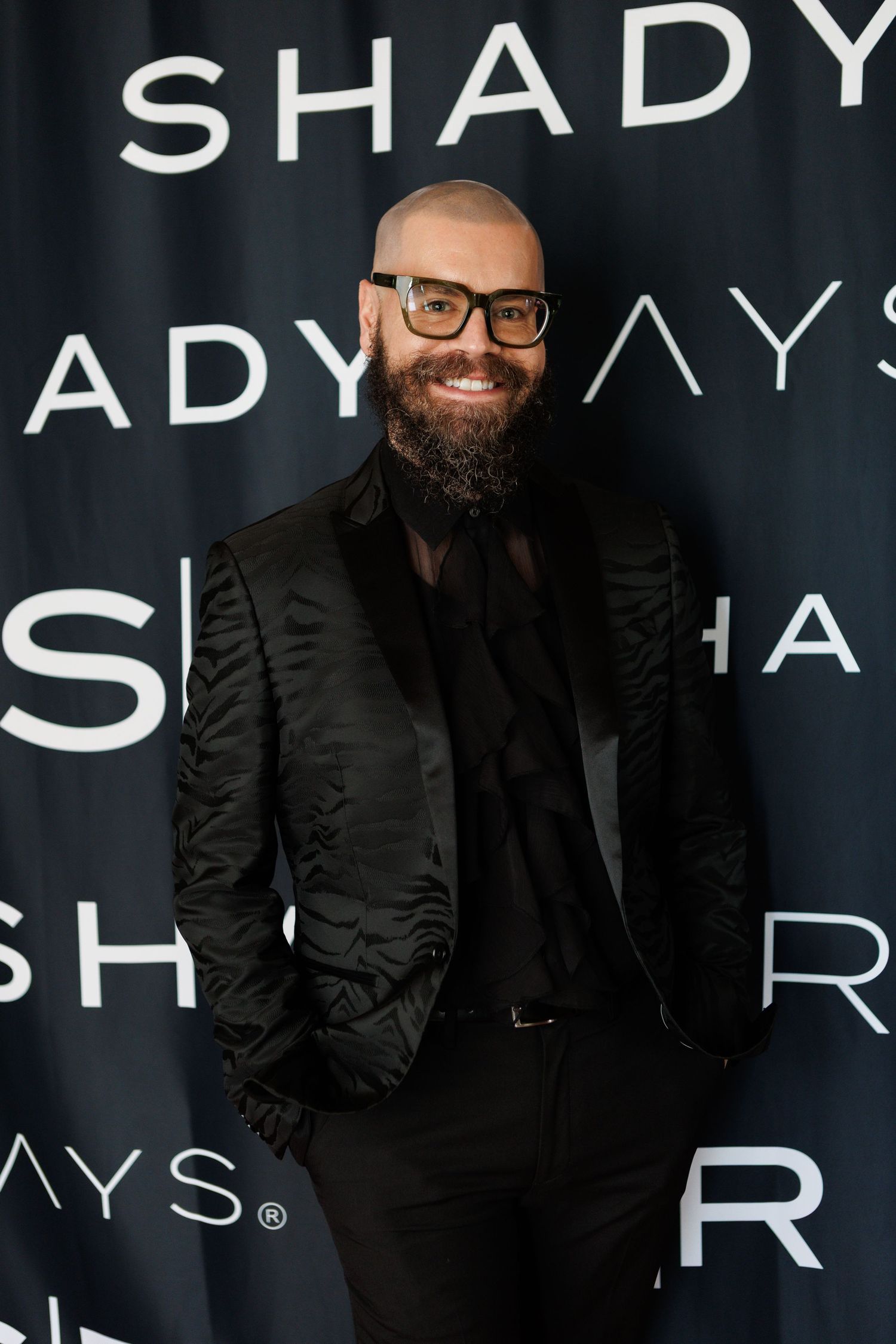Bald bearded man in black tuxedo jacket smiling at Shady Rays event on dark branded backdrop.