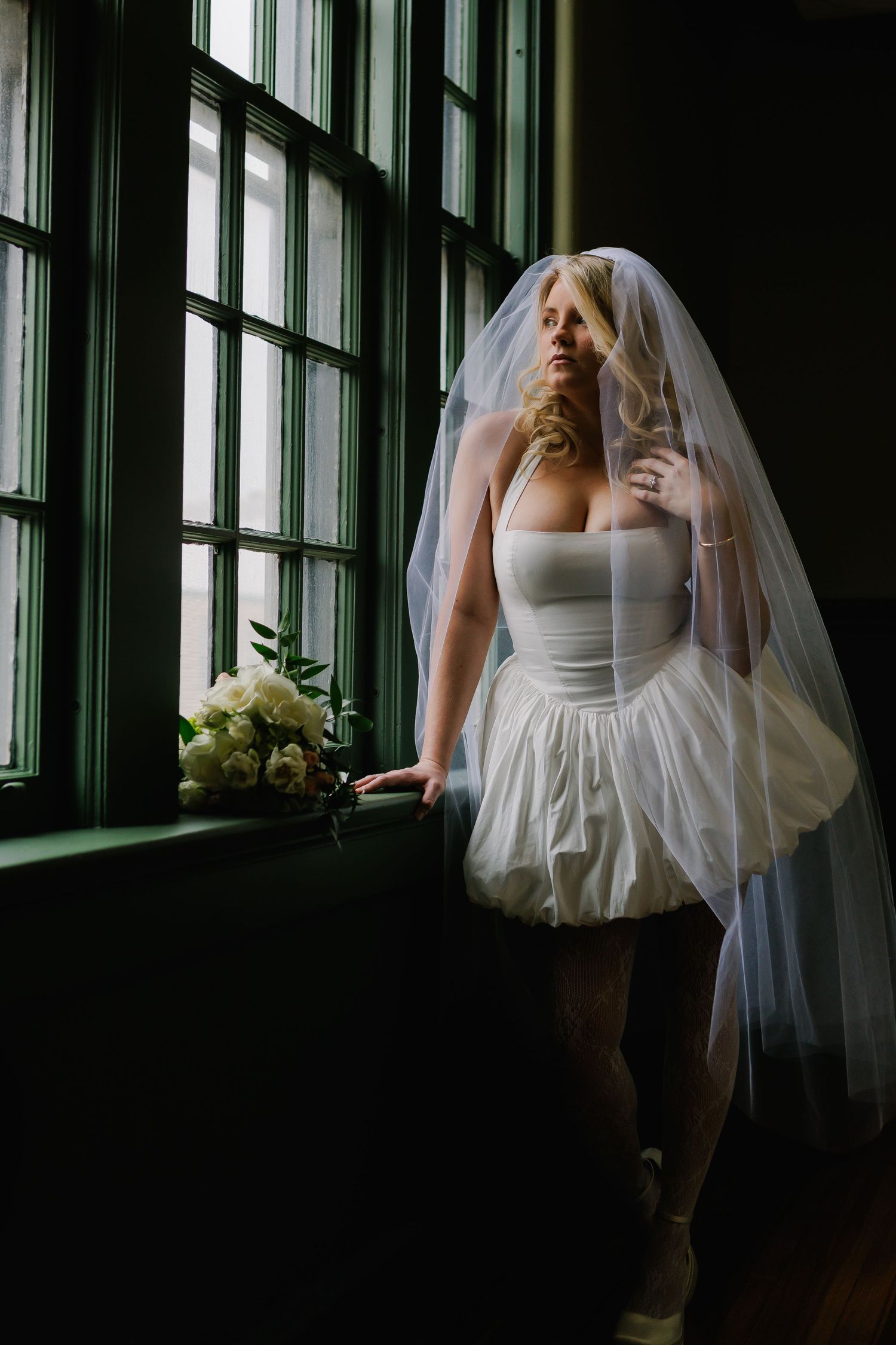 Bride in short white dress and veil stands by dark window with white floral bouquet in moody indoor setting.