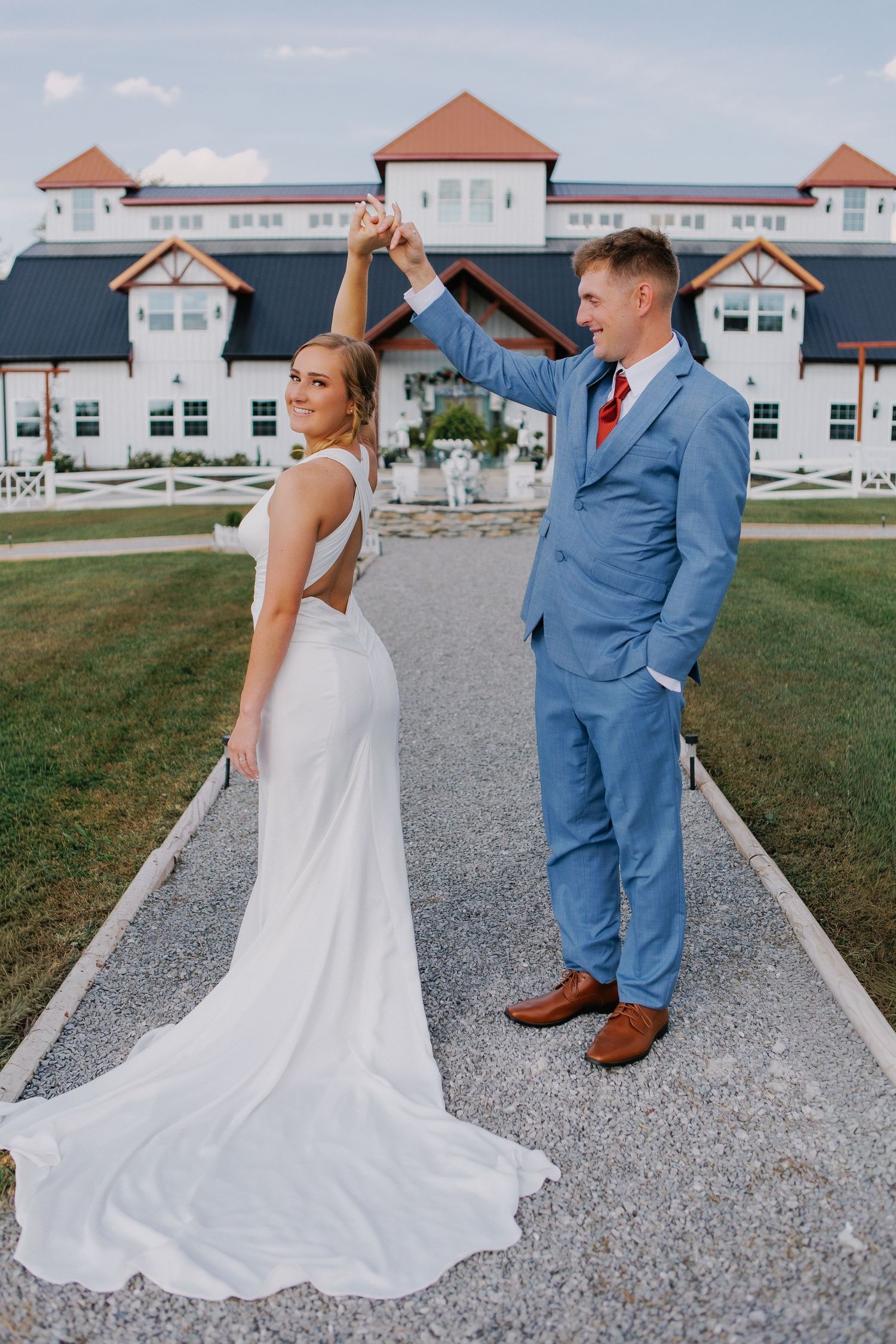 Couple in formal attire pose on path with elegant white building in background.