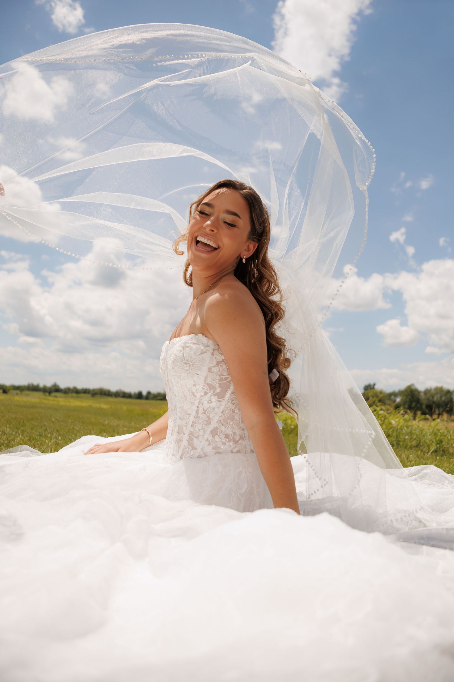 Joyful bride in white lace gown sits outdoors, veil flowing in wind against blue sky and green field.