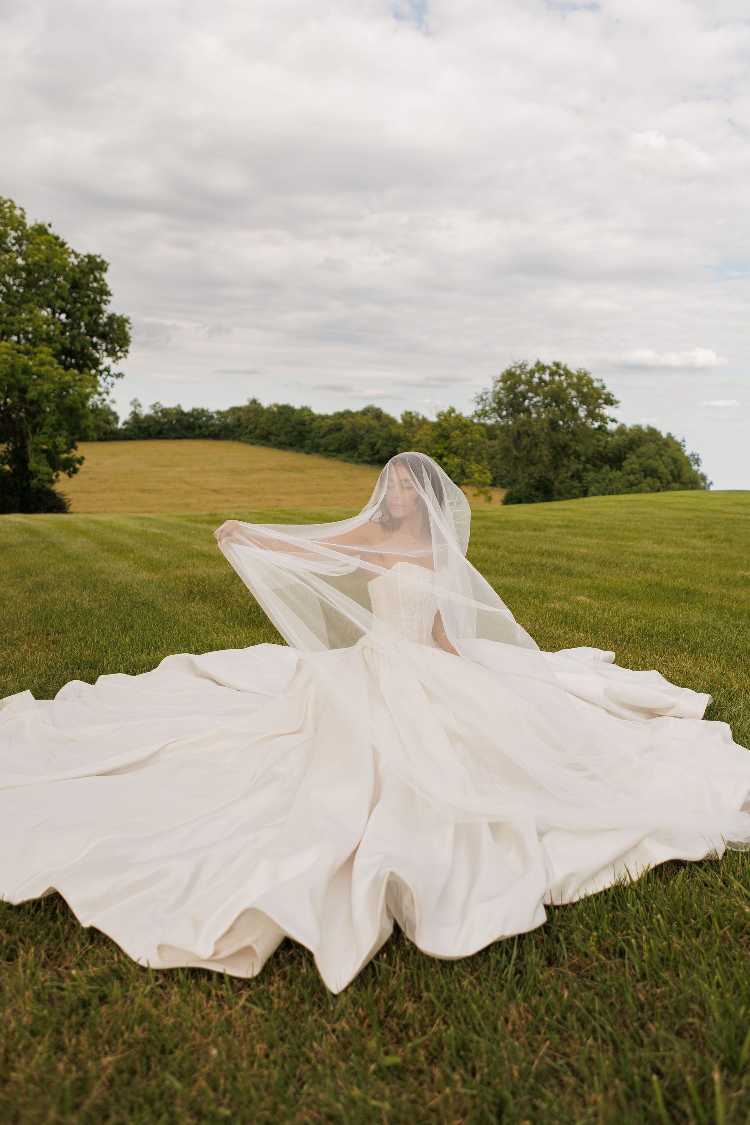 Bride in flowing white gown and veil standing in lush green field with rolling hills and cloudy sky backdrop.